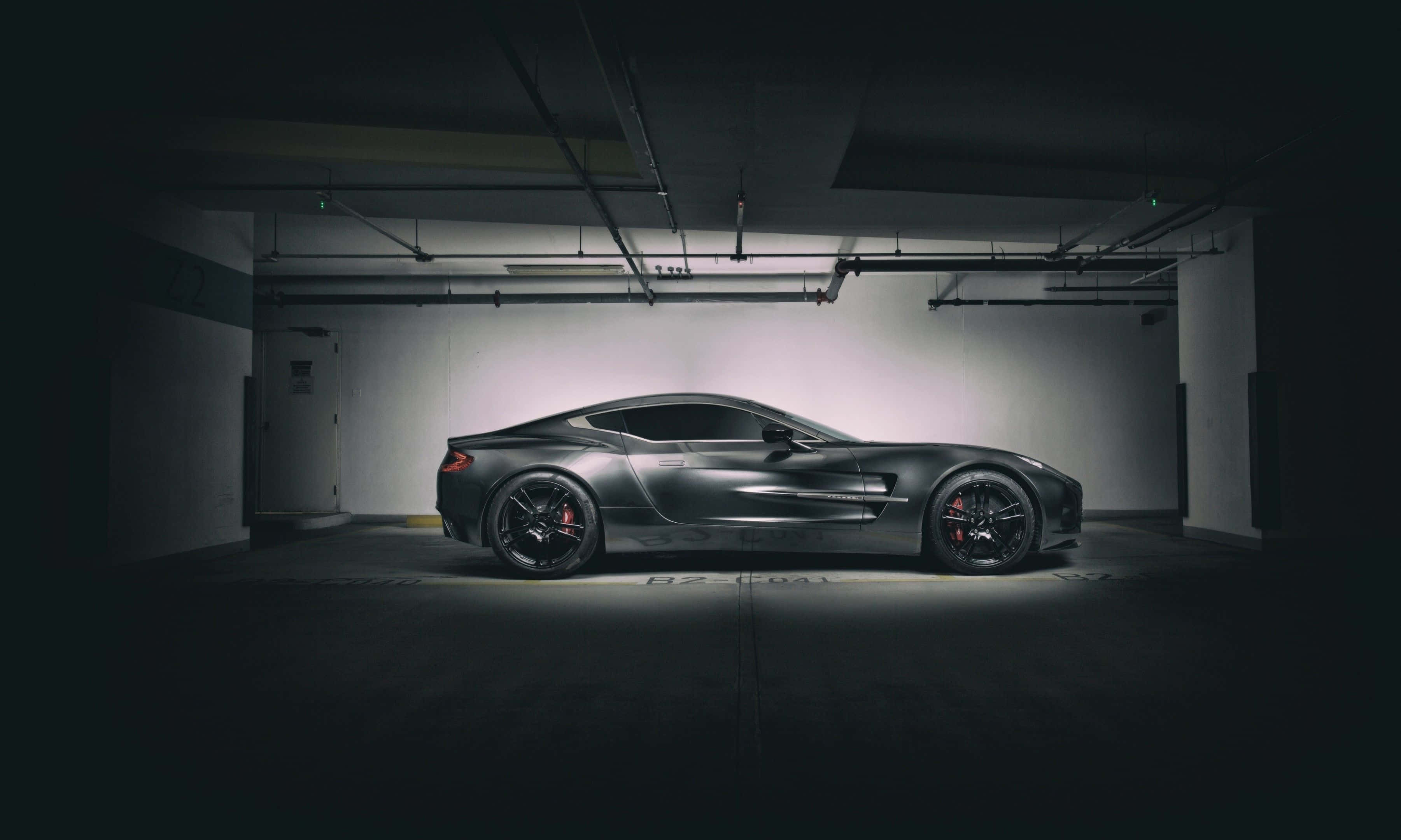 Aston Martin One-77 桌布 壁紙 — 一輛時尚的啞光灰色跑車配備紅色制動卡鉗，在昏暗的地下停車場中被戲劇性地照亮。
