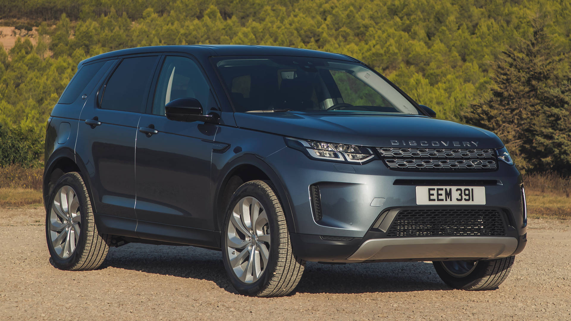 land rover iPhone 桌布 壁紙 — 一輛時尚的啞光藍色 Land Rover Discovery 停在碎石路上，背景是茂密的綠樹，在明亮的日光下。