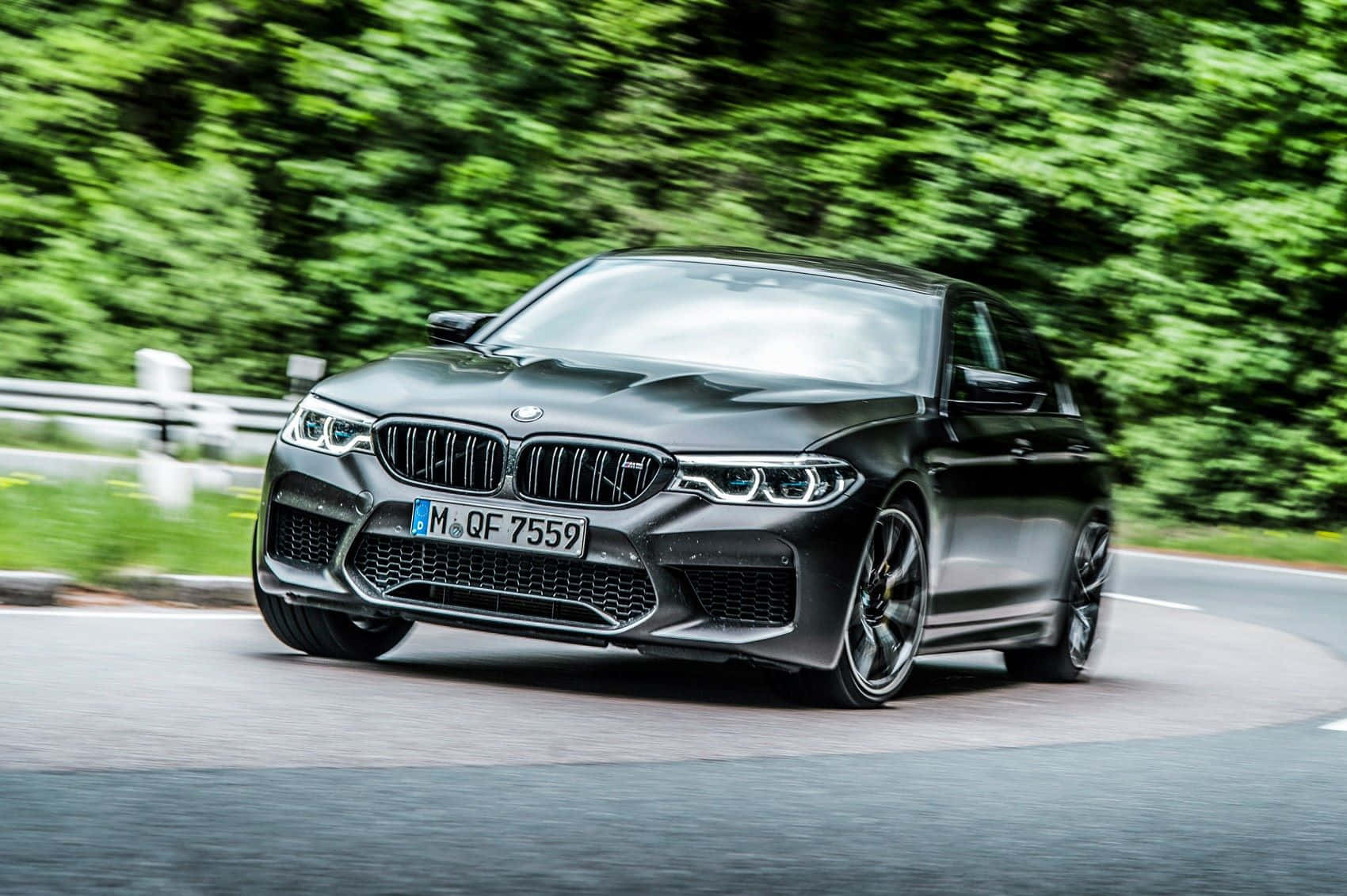 Hybrid Car 桌布 壁紙 — 一輛時尚的啞光黑色BMW M5在蜿蜒的道路上疾馳，周圍環繞著茂密的綠樹。