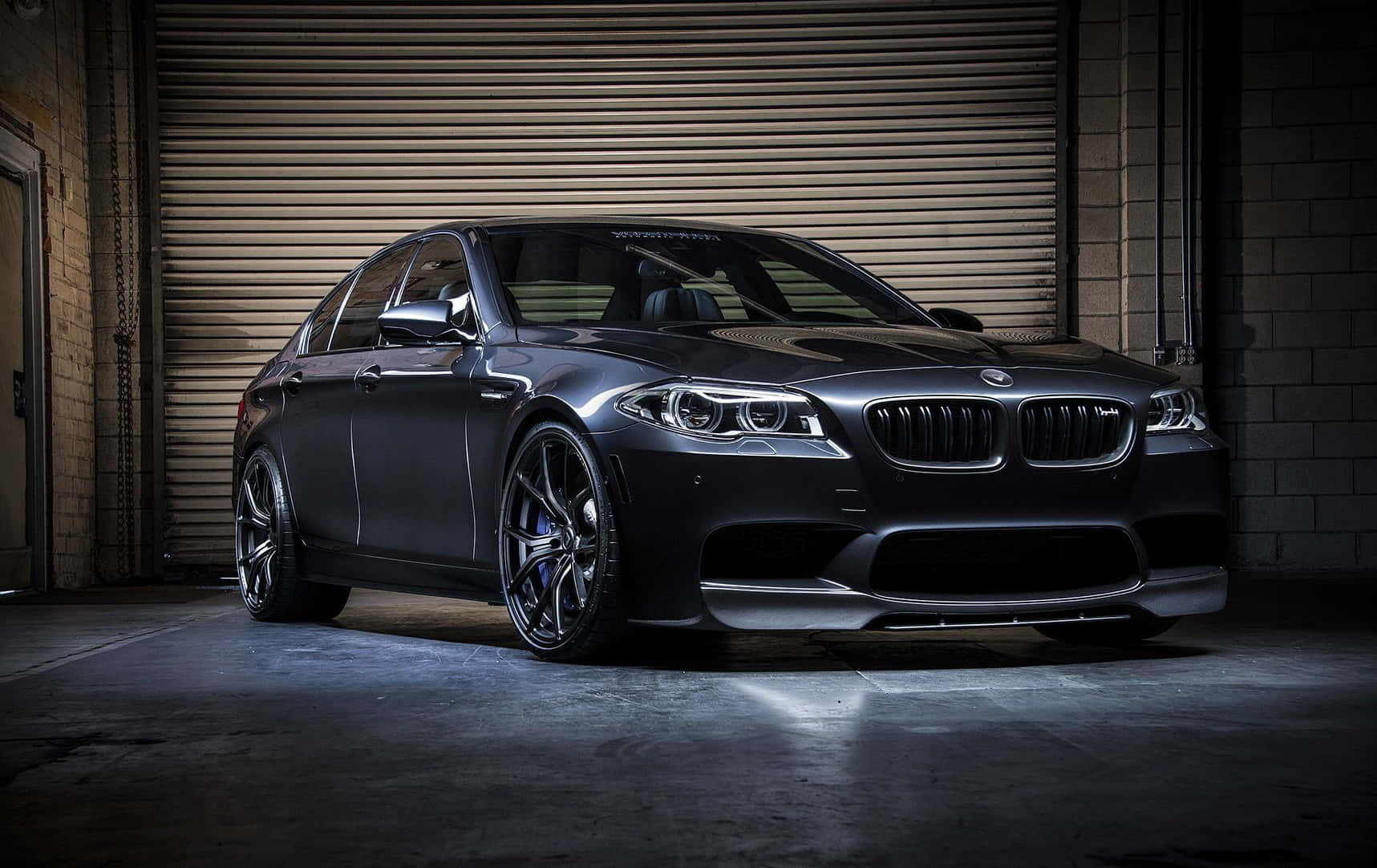 Bmw M5 桌布 壁紙 — 一輛時尚的啞光黑色BMW M5運動轎車停放在光線昏暗的工業車庫中，車庫有混凝土地板和金屬捲門。