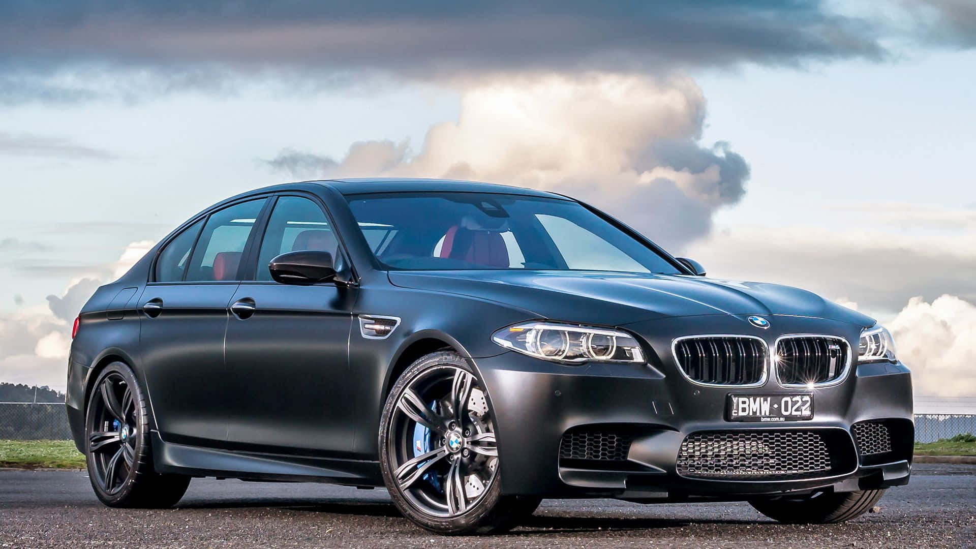 Bmw M5 桌布 壁紙 — 一輛時尚的啞光黑色BMW M5運動轎車停在瀝青路上，背景是戲劇性的多雲天空，車牌清晰可見。