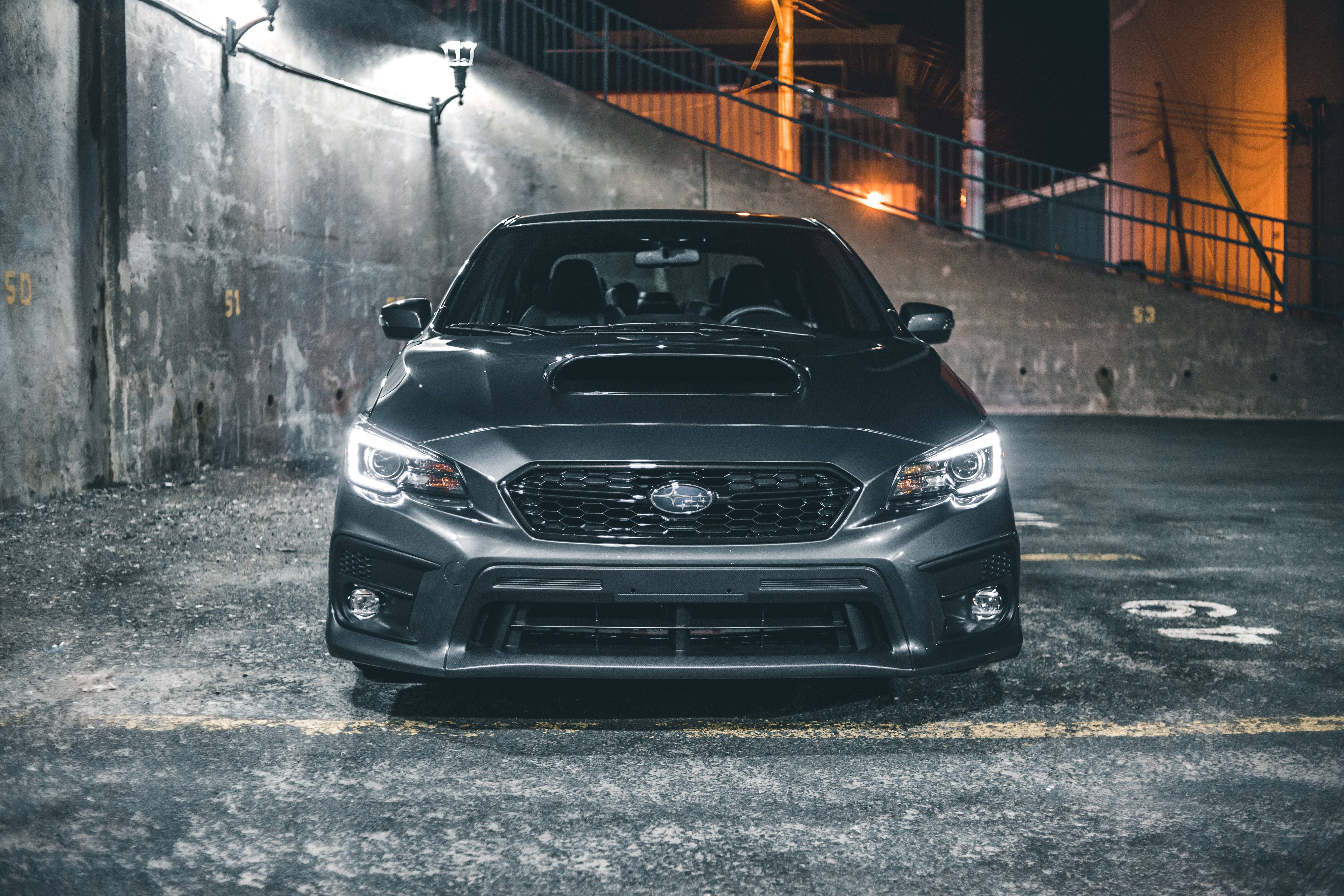 昴 桌布 壁紙 — 一輛時尚的啞光黑色Subaru WRX STI停放在燈光昏暗的市區停車場，車頭燈閃爍發光。