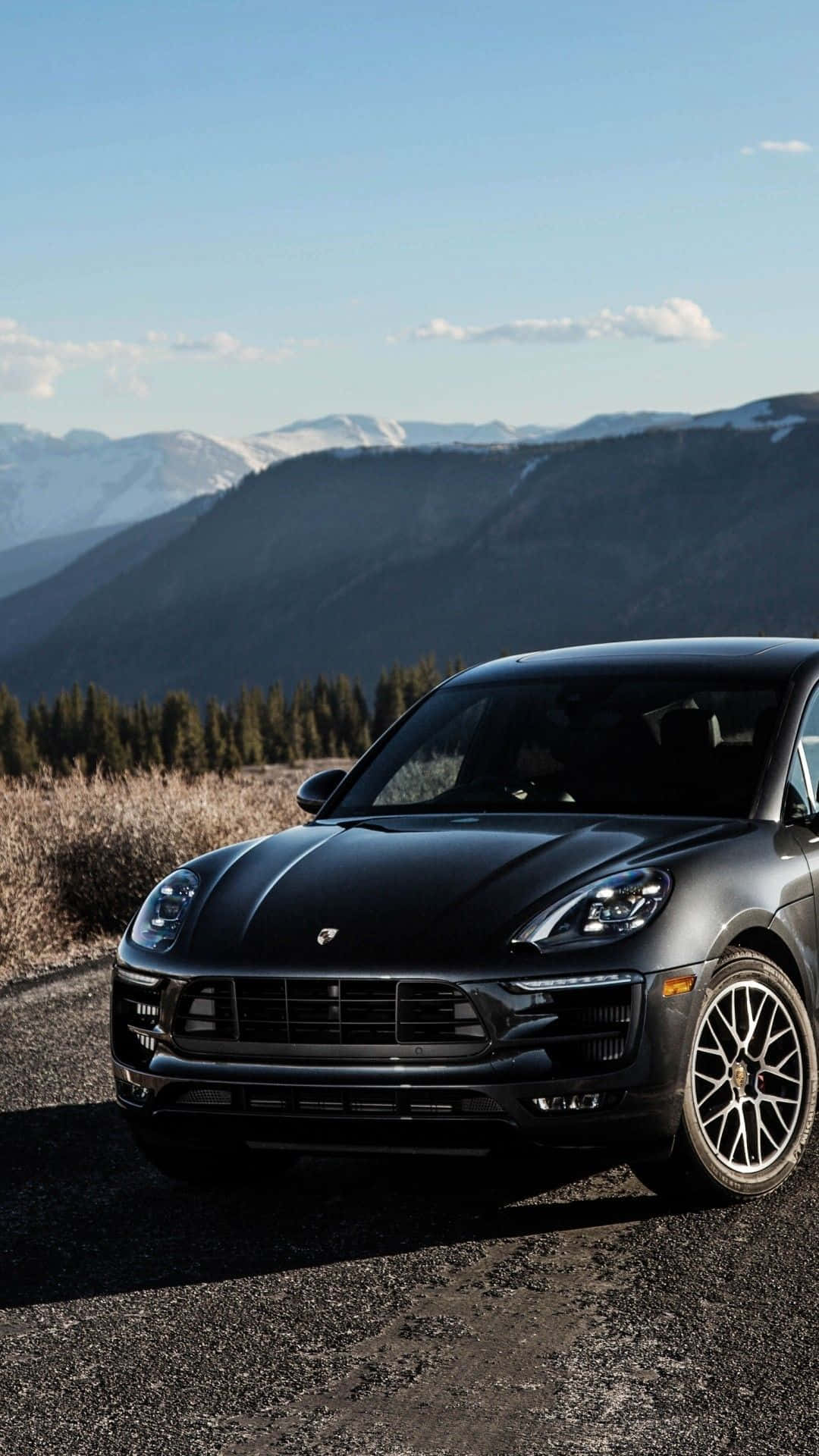 Porsche iPhone 桌布 壁紙 — 一輛時尚的啞光黑色保時捷 Macan 停在碎石路上，背景是白雪覆蓋的山脈和常青樹，天空清澈湛藍。