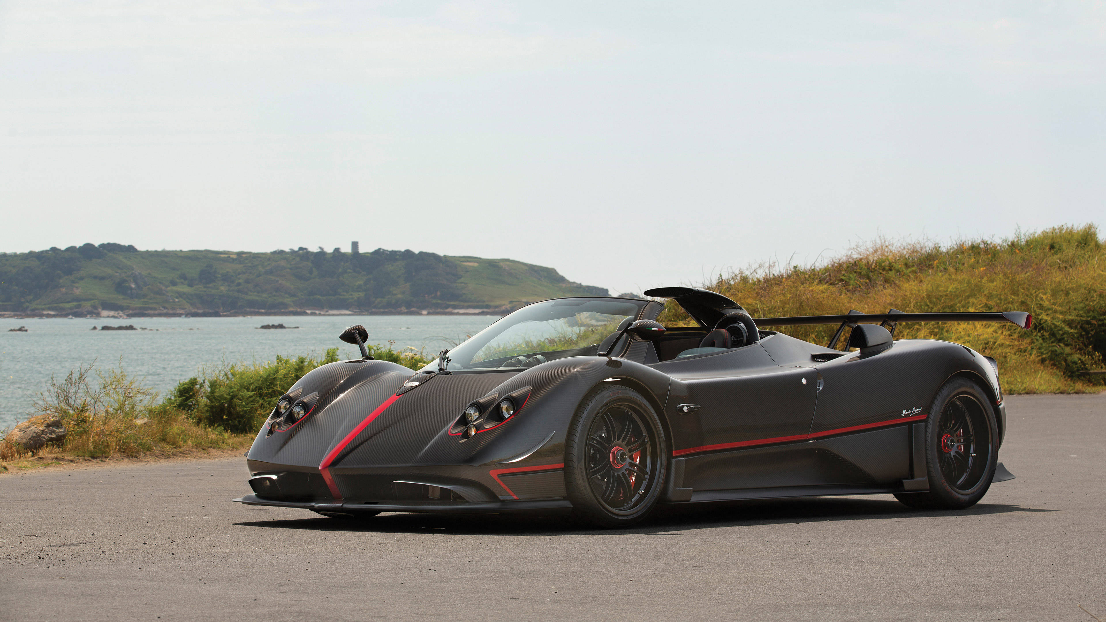 pagani iPhone 桌布 壁紙 — 一輛時尚的啞光黑色帕加尼Zonda敞篷跑車，帶有紅色點綴，停泊在俯瞰平靜大海和綠色山丘的海岸公路上。