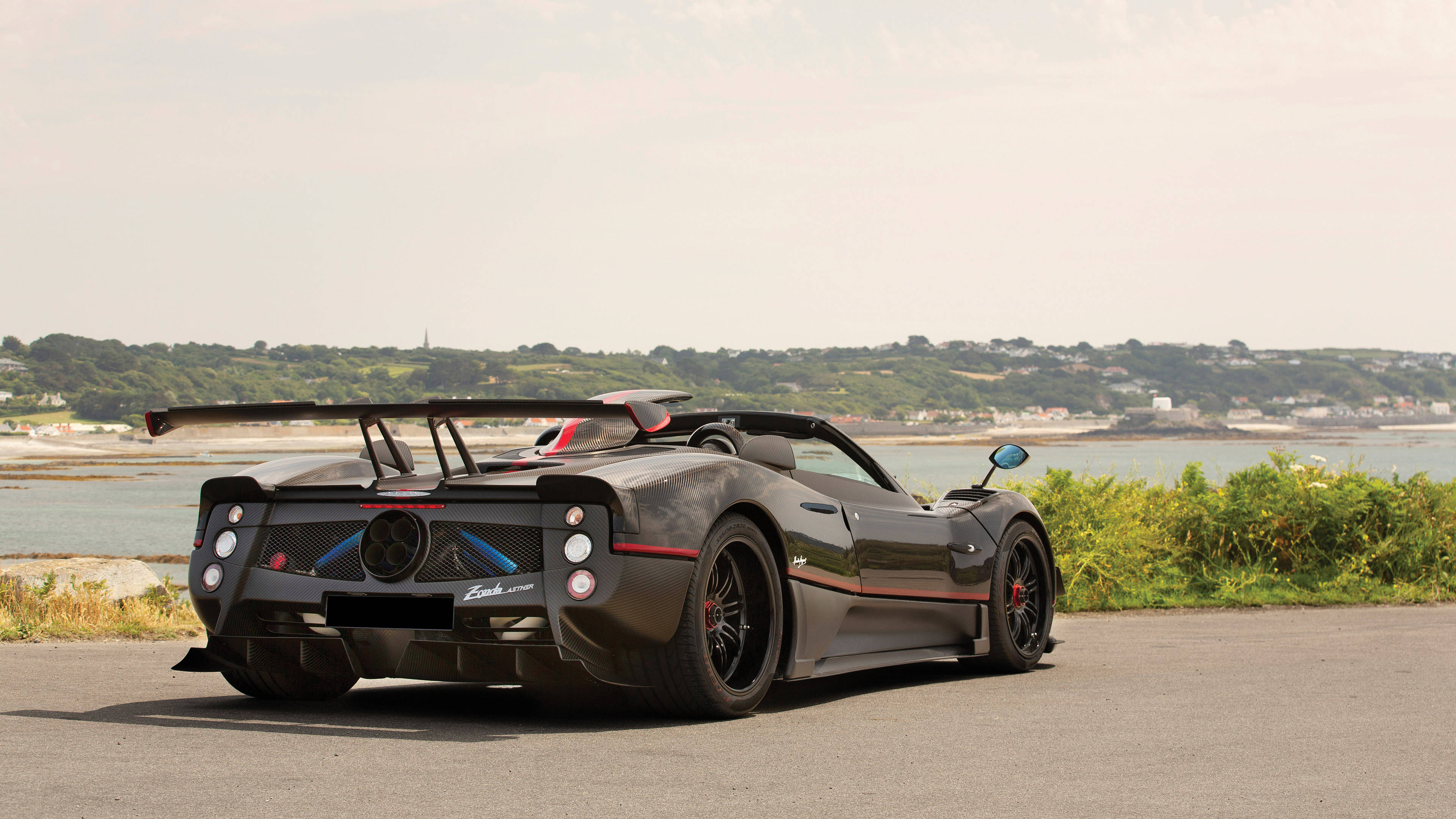 pagani iPhone 桌布 壁紙 — 一輛時尚的啞光黑色帕加尼Zonda跑車帶有紅色點綴，停泊在沿海公路上，俯瞰平靜的海面和綠色山丘，天空朦朧。
