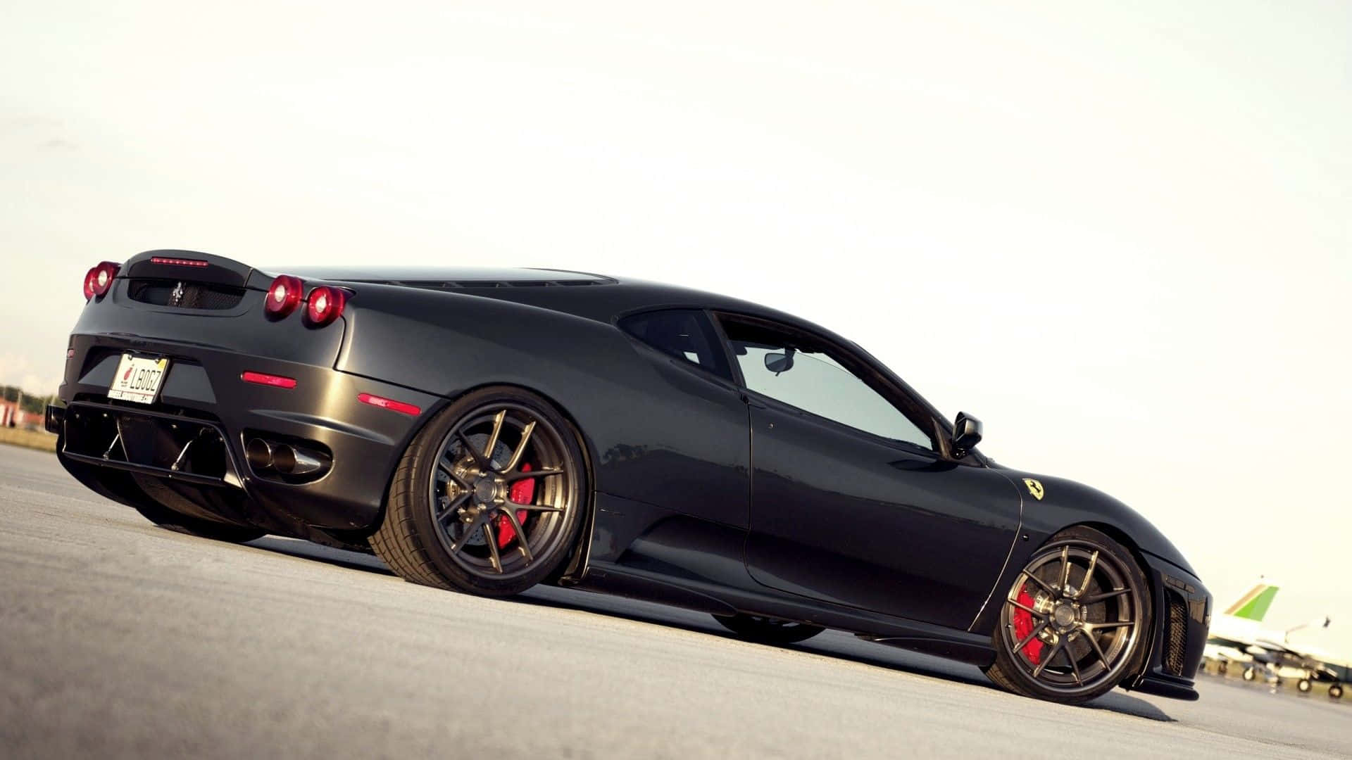 Ferrari F430 桌布 壁紙 — 一輛時尚的啞光黑色法拉利跑車，配備紅色制動卡鉗，在明亮的日光下停放在機場停機坪上。