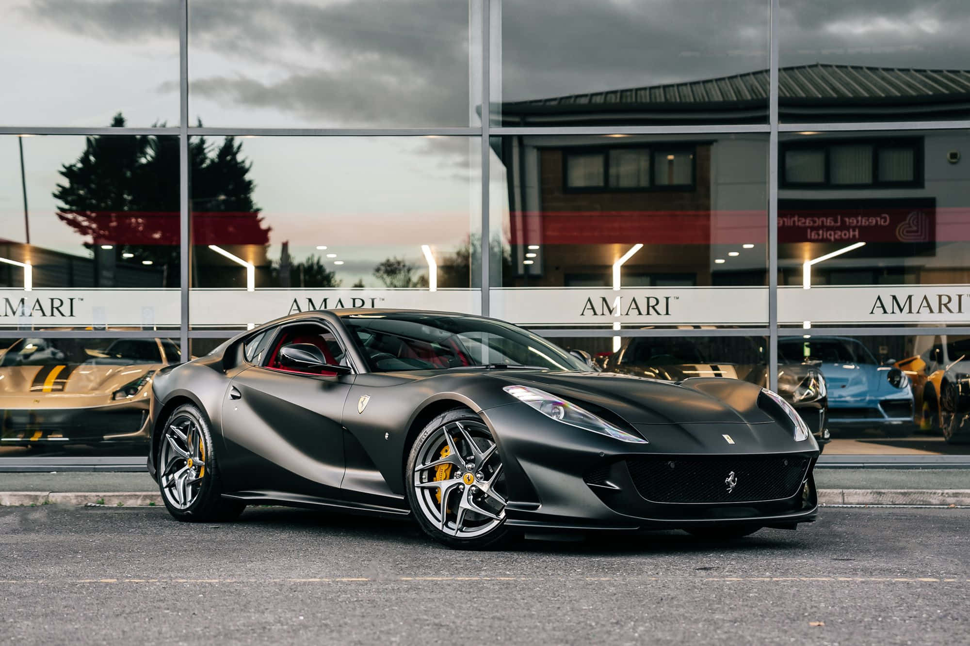 Ferrari 812 Superfast 桌布 壁紙 — 一輛時尚的啞光黑色法拉利跑車停在一家現代玻璃經銷商外，內部可見其他豪華車。