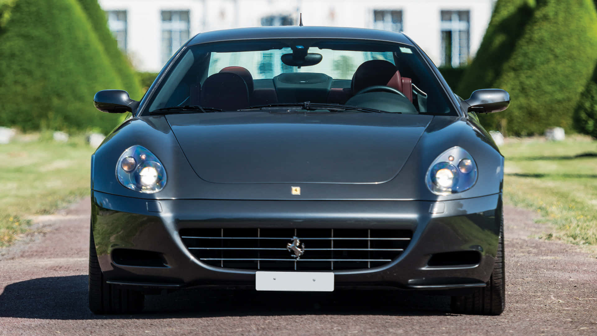 Ferrari 612 Scaglietti 桌布 壁紙 — 一輛時尚的啞光黑色法拉利跑車停在碎石車道上，背景是修剪整齊的樹籬和白色建築。