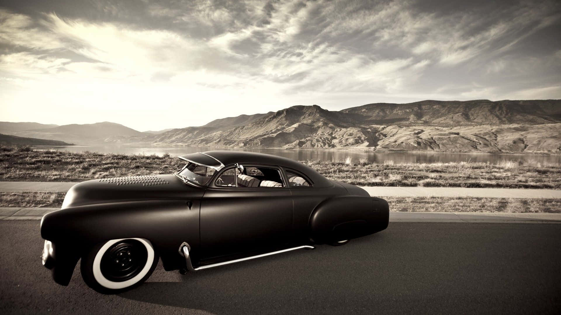 Hot Rod 桌布 壁紙 — 一輛時尚的啞光黑色經典汽車在沙漠公路上行駛，背景有山脈和湖泊，天空烏雲密佈，氣氛戲劇化。