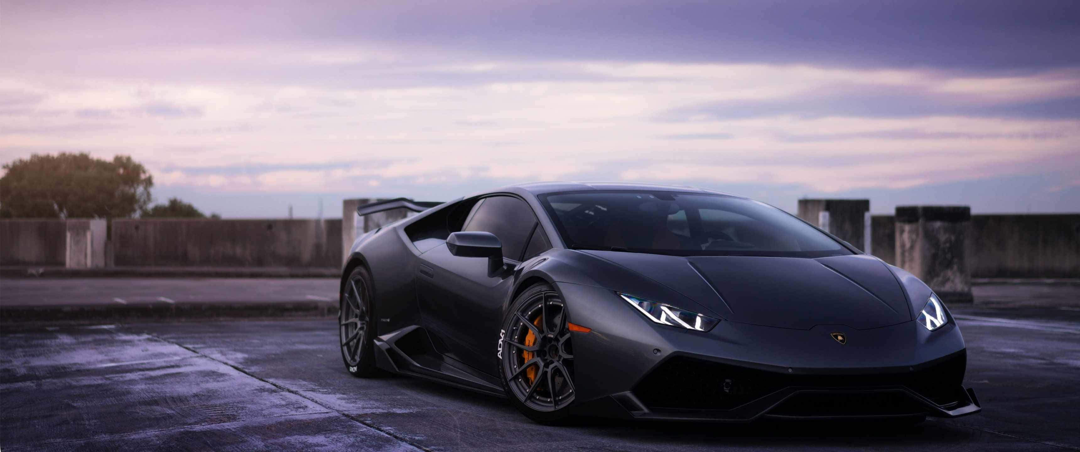3440x1440p Lamborghini 桌布 壁紙 — 一輛時尚的啞光黑色藍寶堅尼Huracán跑車停放在潮濕的屋頂上，黃昏時分天空多雲。