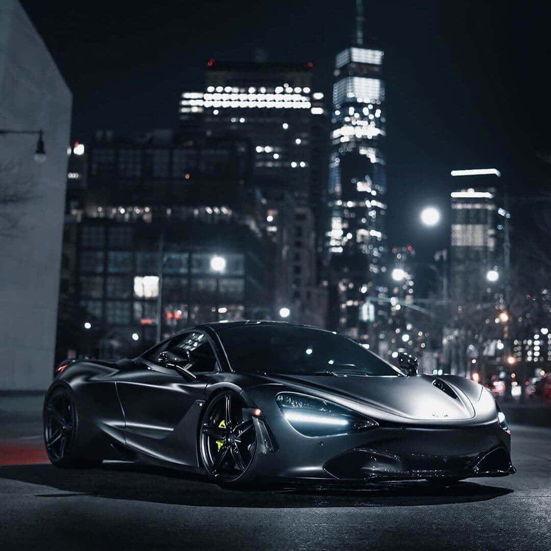 Best Mclaren 720s 桌布 壁紙 — 一輛時尚的啞光黑色跑車在夜晚的城市燈光下閃耀，背景是高聳的摩天大樓。
