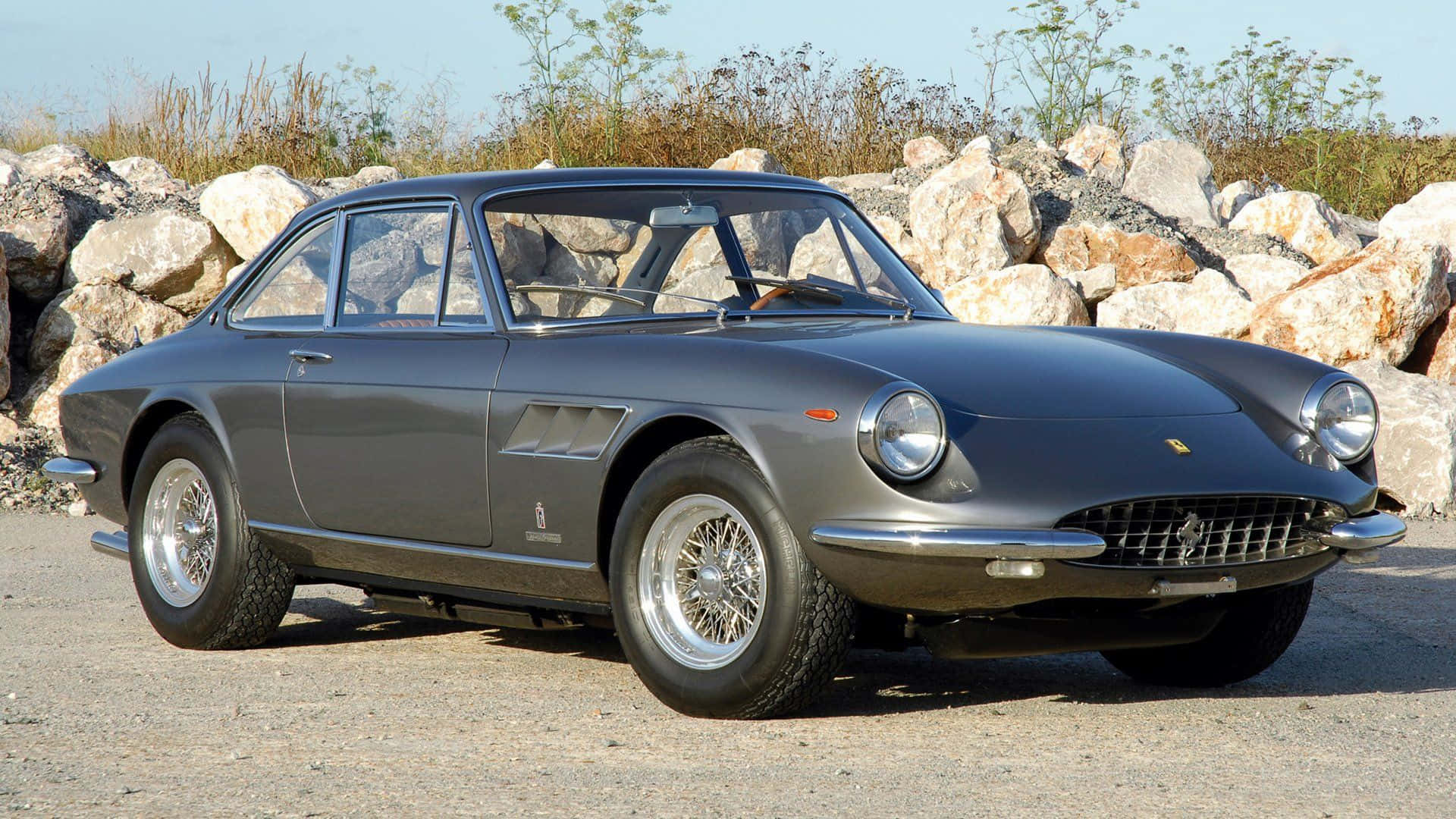 Vintage Ferrari 桌布 壁紙 — 一輛時尚的復古灰色法拉利跑車停泊在碎石上，旁邊是岩石露頭，天空晴朗蔚藍。