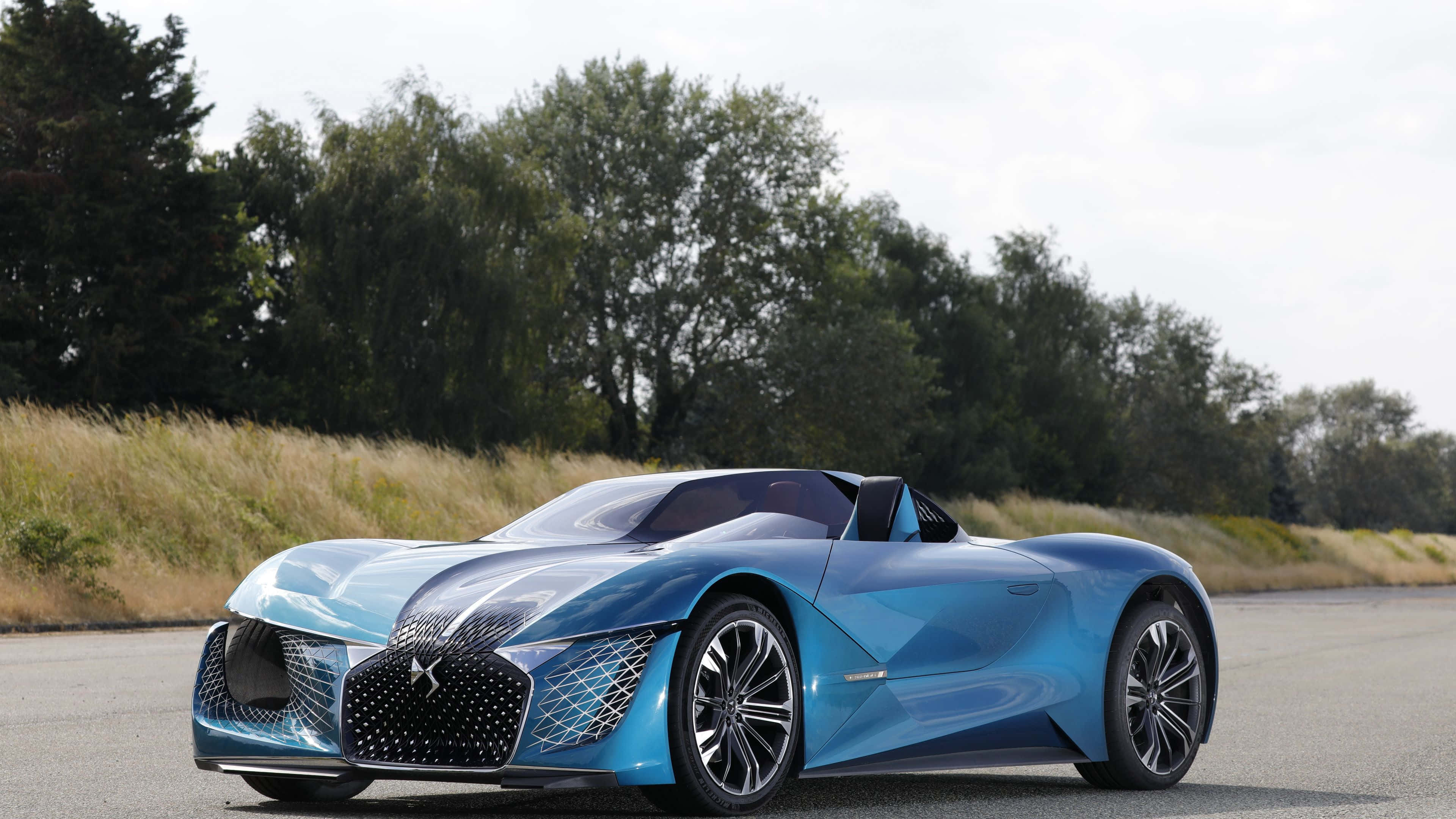Ds Automobiles 桌布 壁紙 — 一輛時尚的未來主義藍色敞篷跑車，在陰天中行駛在一條鋪砌的道路上，兩旁是高高的草叢和茂密的樹木。