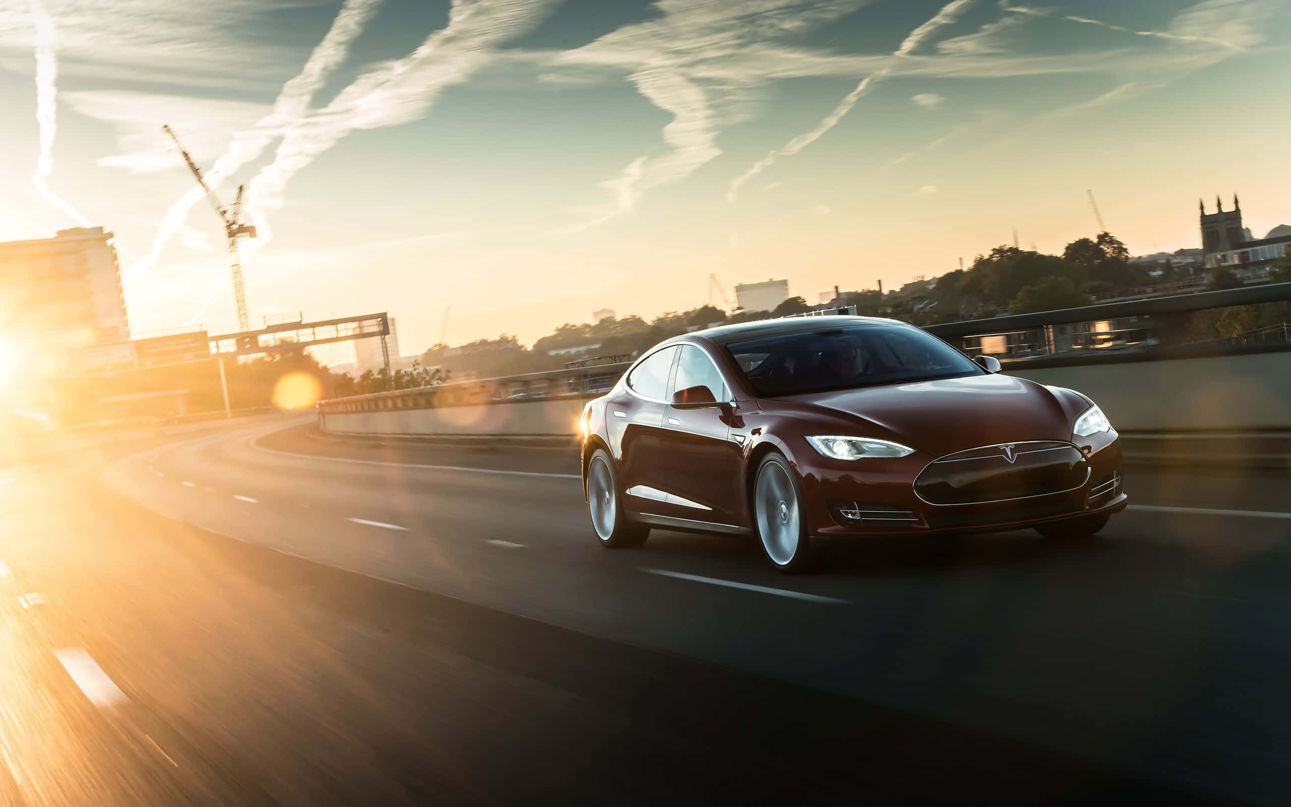 特斯拉 桌布 壁紙 — 一輛時尚的栗色Tesla Model S在金色日落時分在高速公路上巡航，背景是城市天際線和起重機。