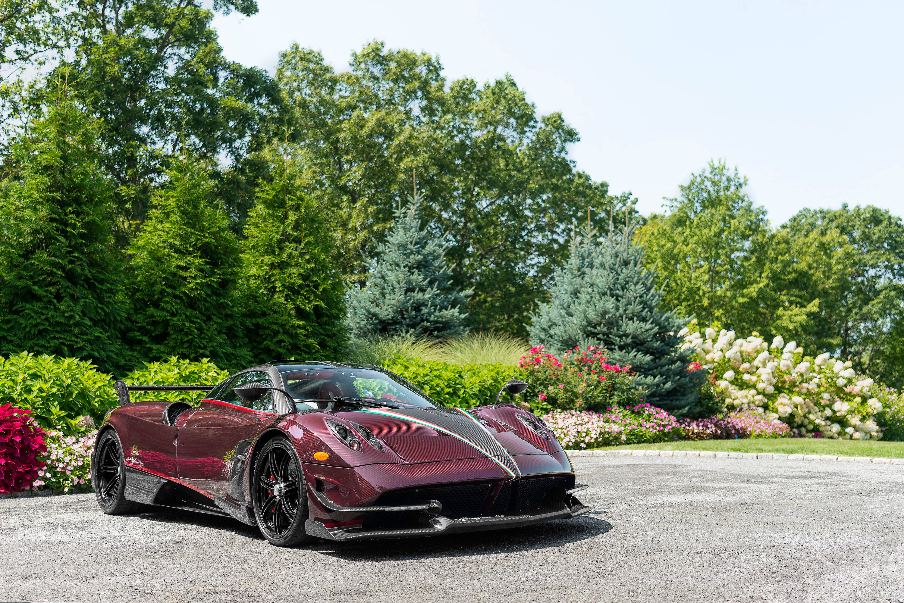 Pagani 桌布 壁紙 — 一輛時尚的栗色帕加尼Huayra跑車在明亮的陽光下閃閃發光，停在碎石車道上，周圍環繞著茂密的綠樹和色彩繽紛的花壇。