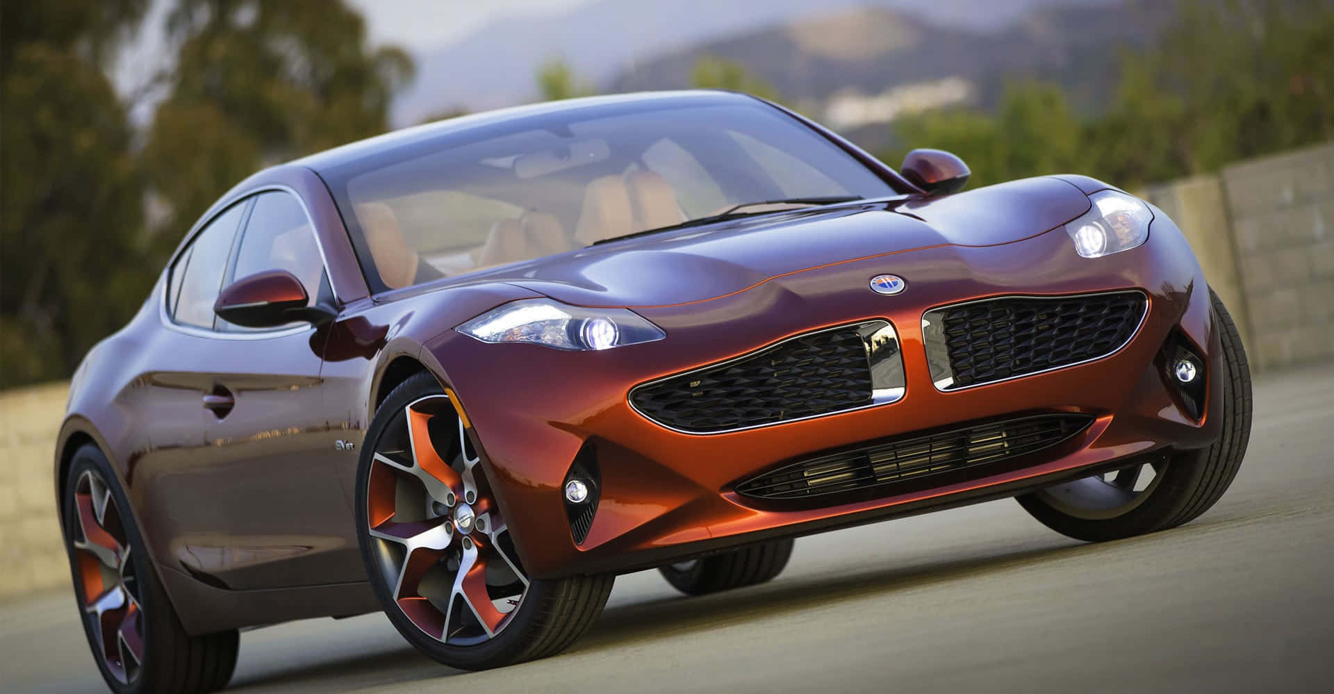 Fisker 桌布 壁紙 — 一輛時尚的栗色跑車，配備橙色輪圈，沿著陽光普照的道路行駛，背景是模糊的綠色山丘。