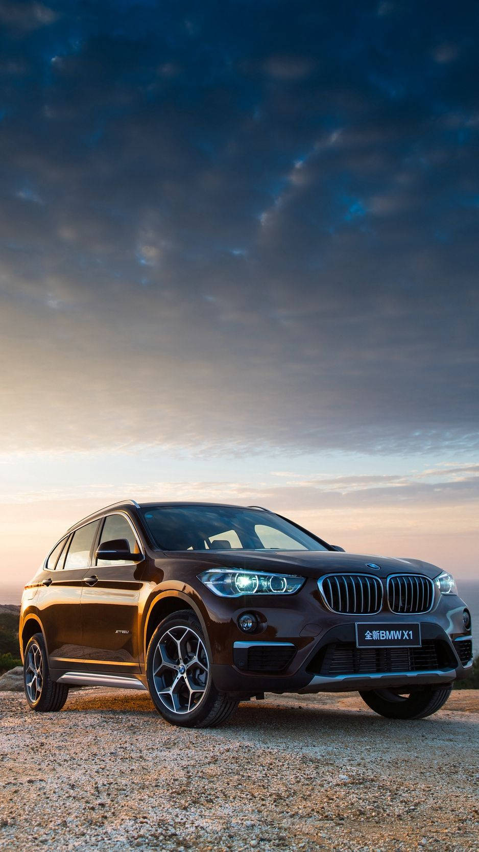 X 桌布 壁紙 — 一輛時尚的棕色BMW X1 SUV停在碎石上，背景是戲劇性的暮色天空，車頭燈閃閃發光。