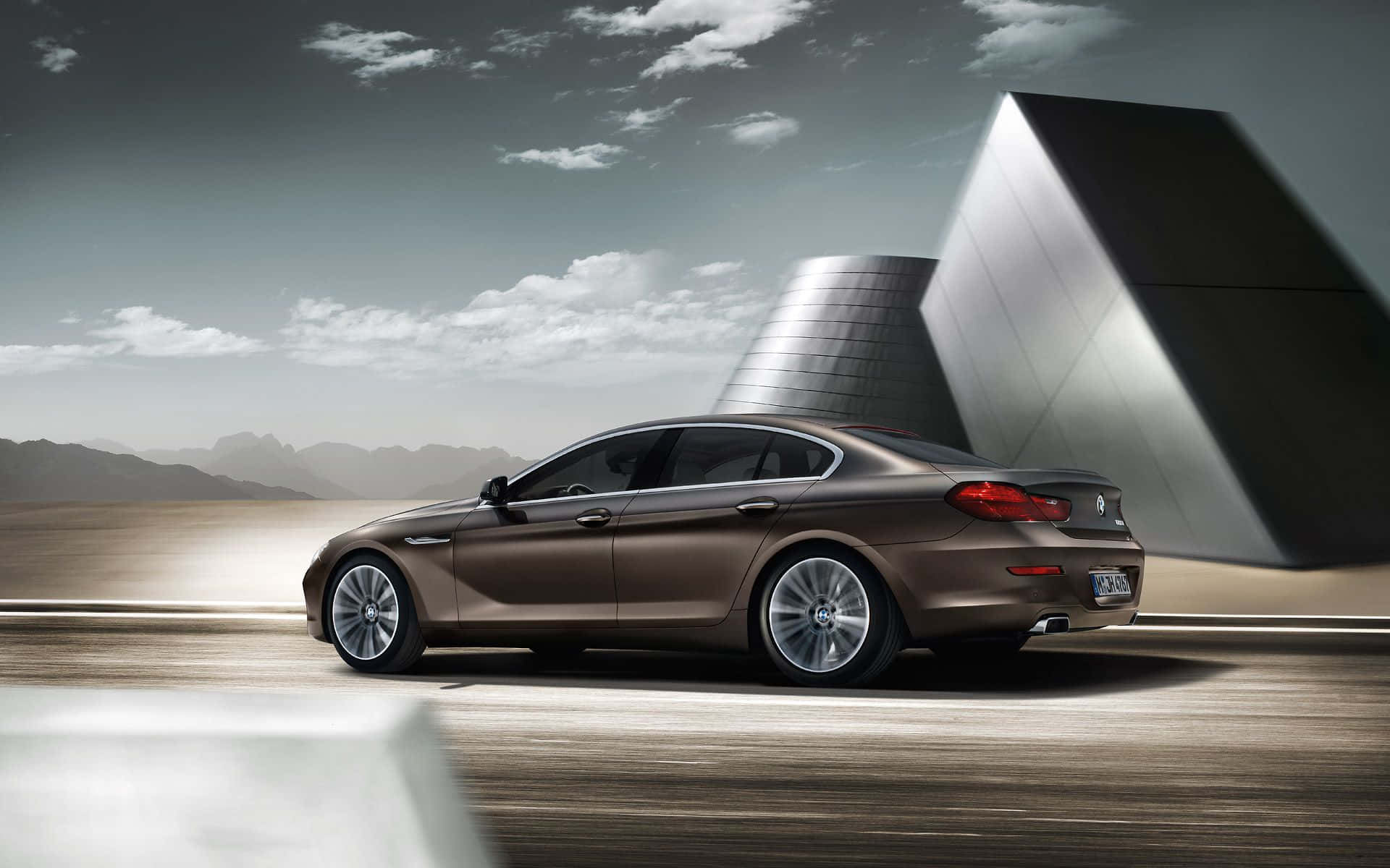Bmw 6 Series 桌布 壁紙 — 一輛時尚的棕色BMW轎車在陰天和遠山的襯托下滑過一座未來主義的金屬建築。