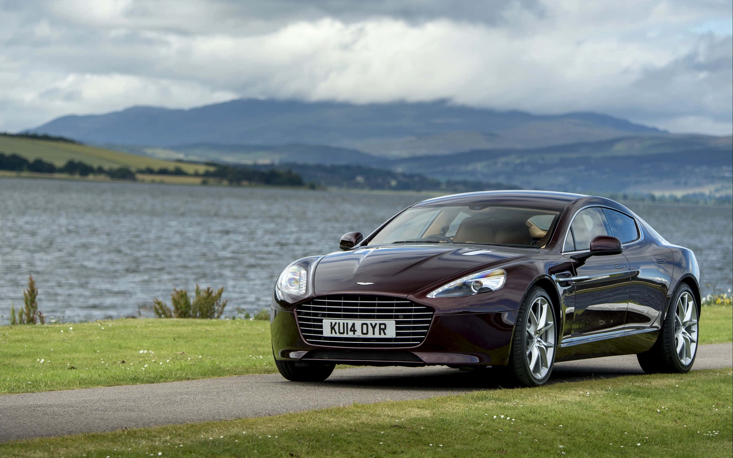 Aston Martin Rapide S 桌布 壁紙 — 一輛時尚的棗紅色Aston Martin跑車停在湖邊的草地上，背景是起伏的山丘和多雲的天空。