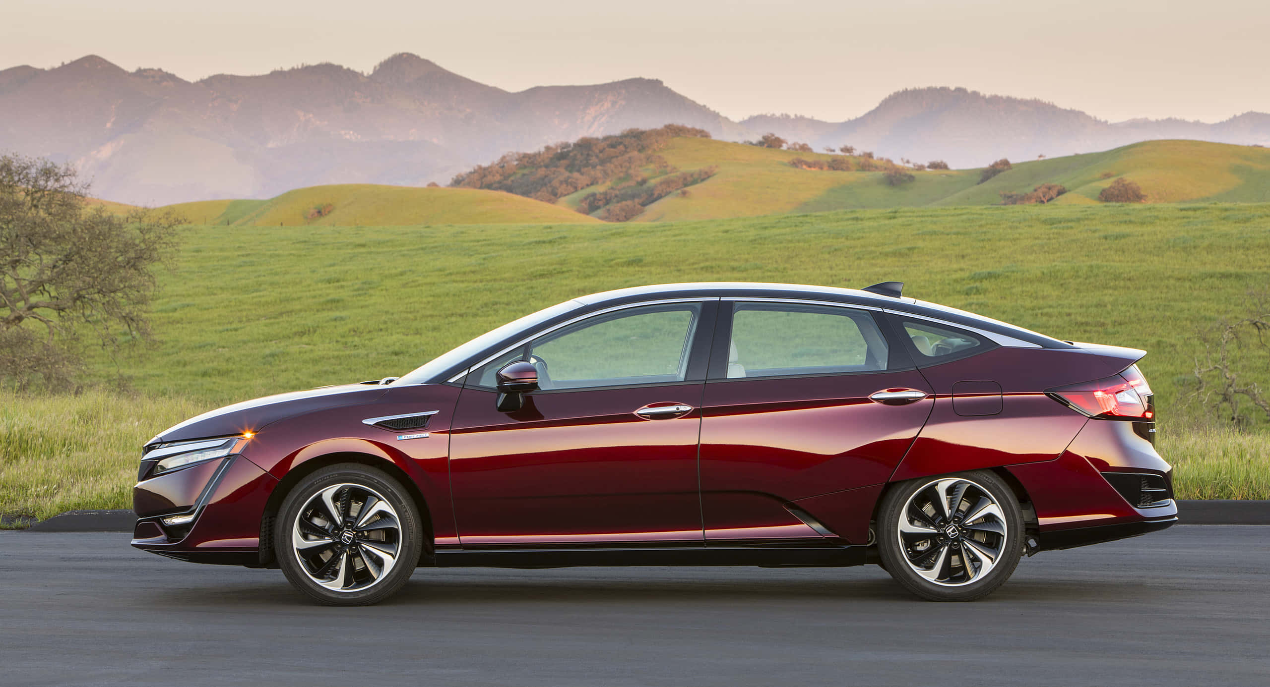 Honda Clarity 桌布 壁紙 — 一輛時尚的棗紅色電動車沿著道路行駛，穿過起伏的綠色山丘，遠處有山脈，在柔和的金色日落天空下。