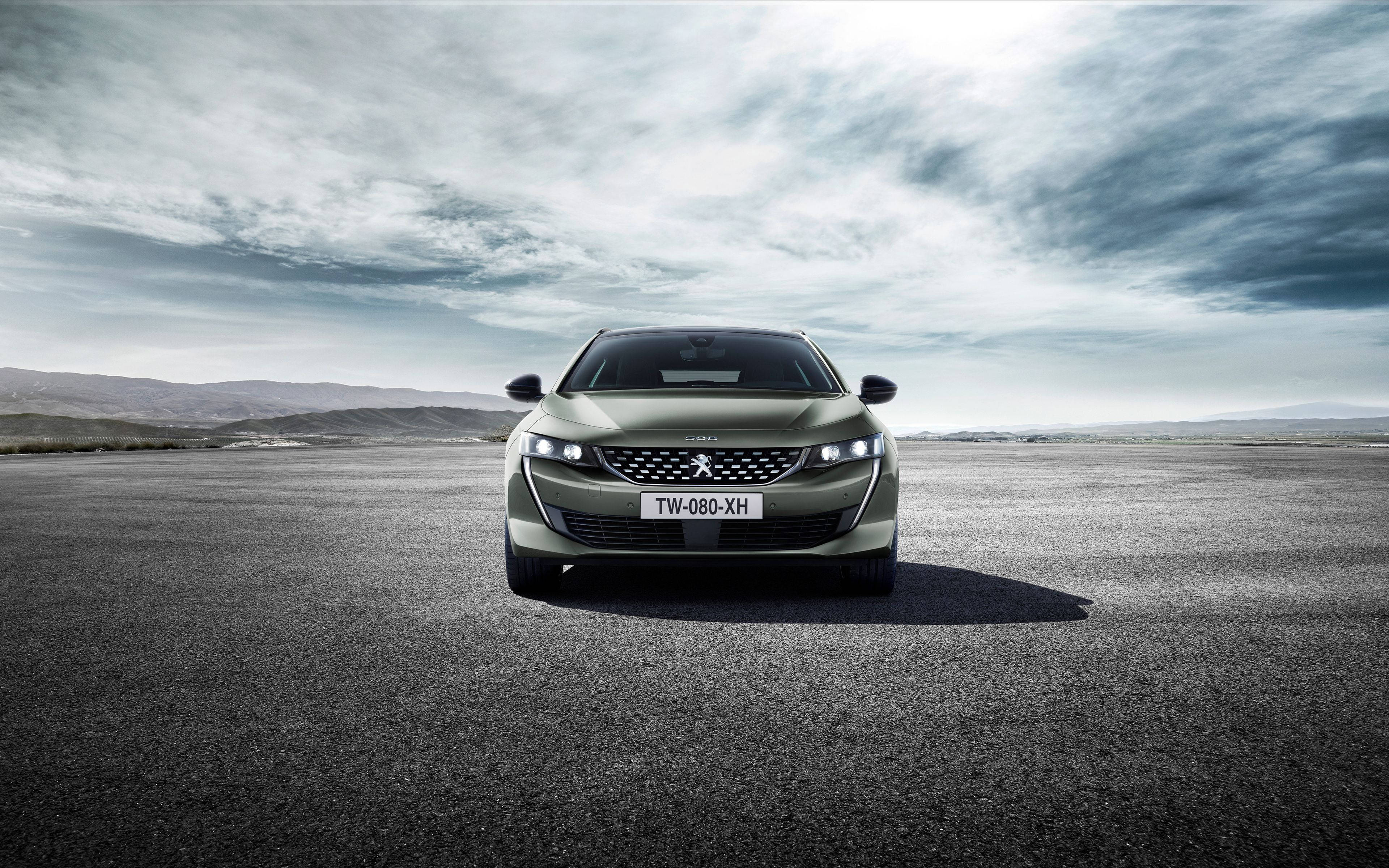 Peugeot 桌布 壁紙 — 一輛時尚的橄欖綠色標緻轎車在廣闊的瀝青平原上向前行駛，背景是戲劇性的多雲天空和遠處的山丘。
