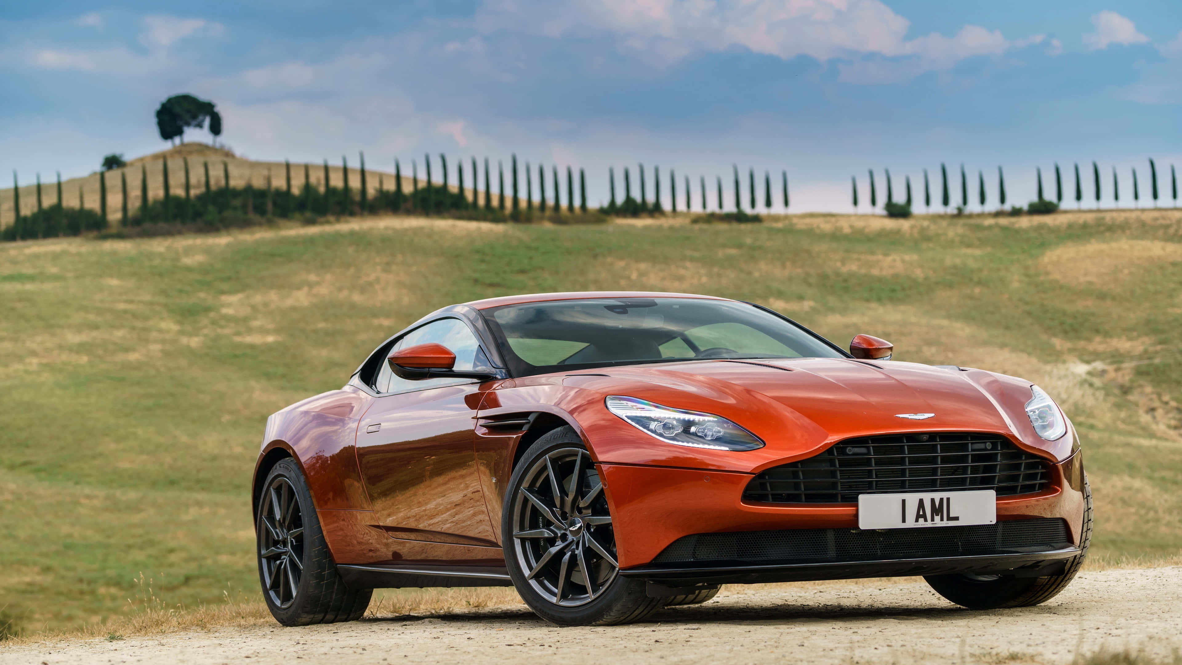 奧斯頓馬丁 桌布 壁紙 — 一輛時尚的橙色Aston Martin跑車停泊在托斯卡尼起伏山丘中的一條泥路上，周圍有柏樹，天空部分多雲。