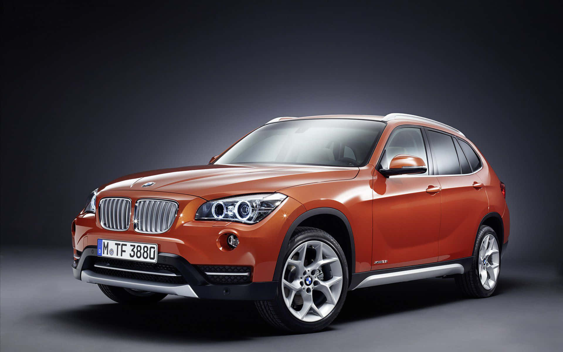 Bmw X1 桌布 壁紙 — 一輛時尚的橙色BMW X1 SUV在攝影棚內以戲劇性燈光和深色漸變背景展示。