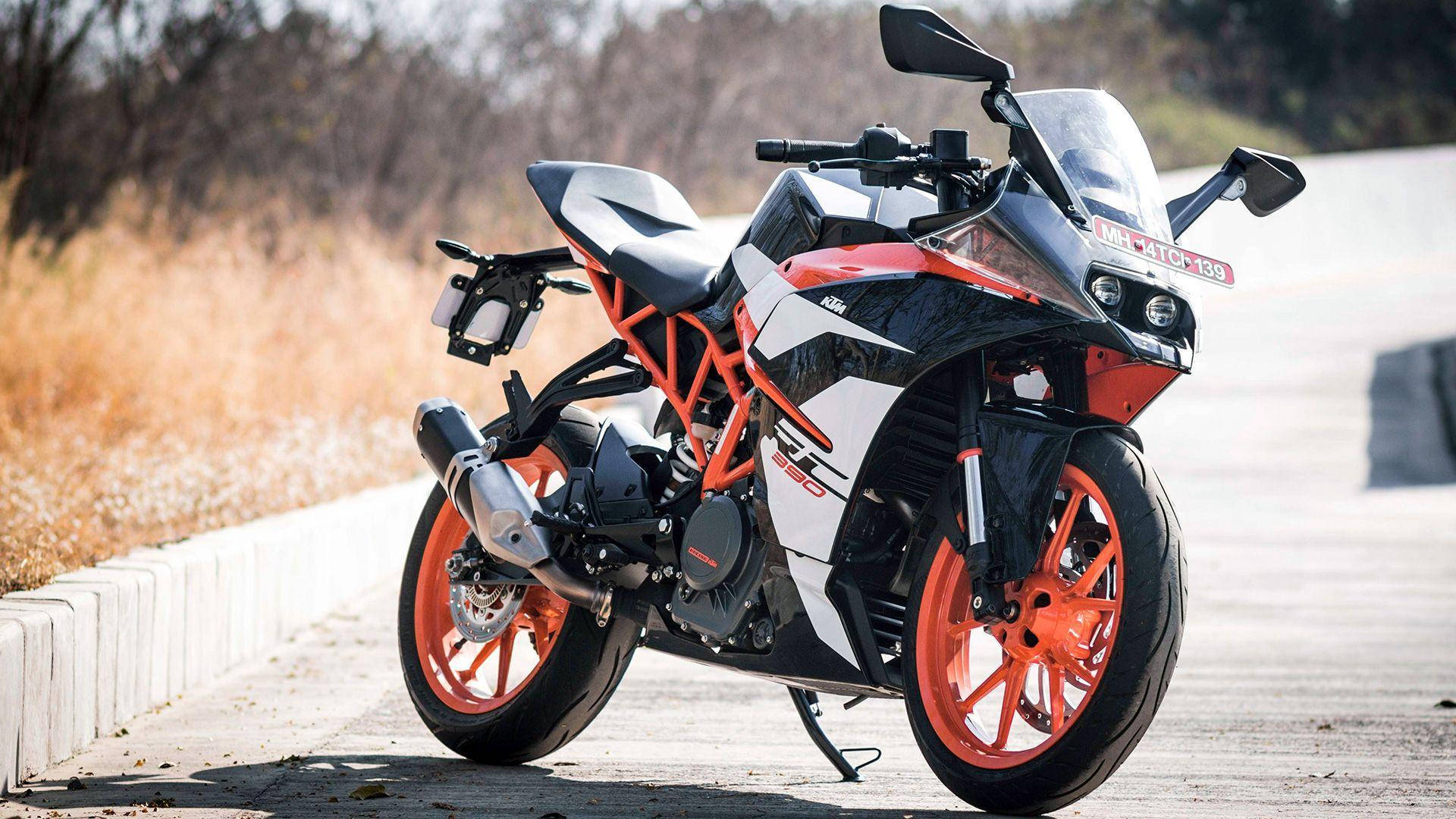 機車 桌布 壁紙 — 一輛時尚的橙色和白色KTM RC 390摩托車停在路邊，有乾草和混凝土路緣，在明亮的陽光下。