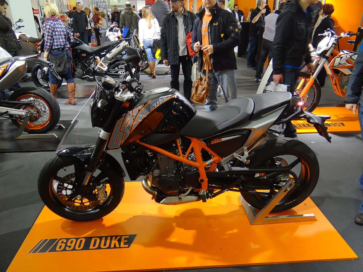 KTM 摩托車 桌布 壁紙 — 一輛時尚的橙色和黑色KTM 690 Duke摩托車在擁擠的室內展覽中展示，與會者走動觀看。