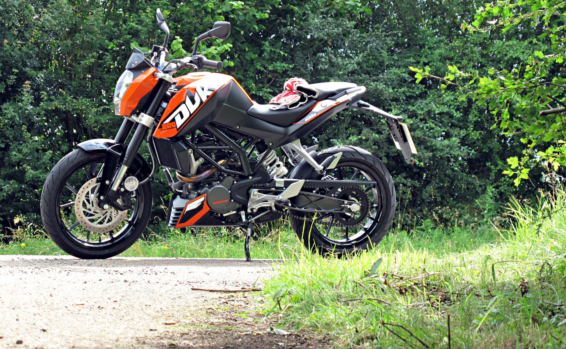  壁紙 — 一輛時尚的橙色和黑色KTM Duke摩托車停在碎石路上，旁邊是茂密的綠色樹葉。