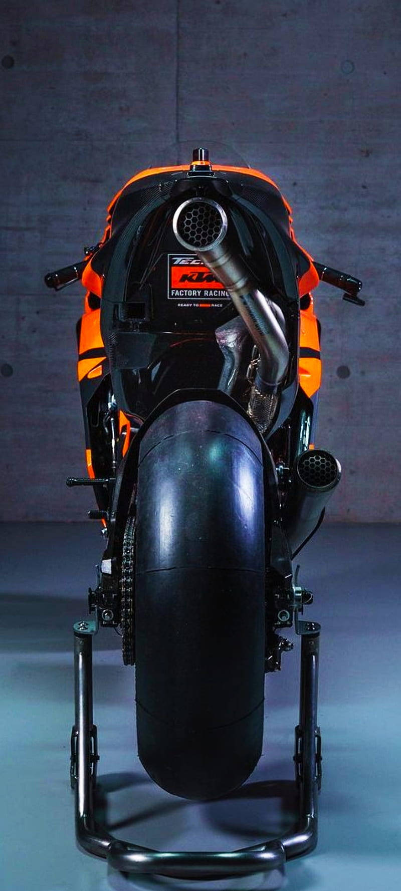 機車 桌布 壁紙 — 一輛時尚的橙色和黑色KTM Factory Racing摩托車放在支架上，背景是混凝土牆的後視圖。