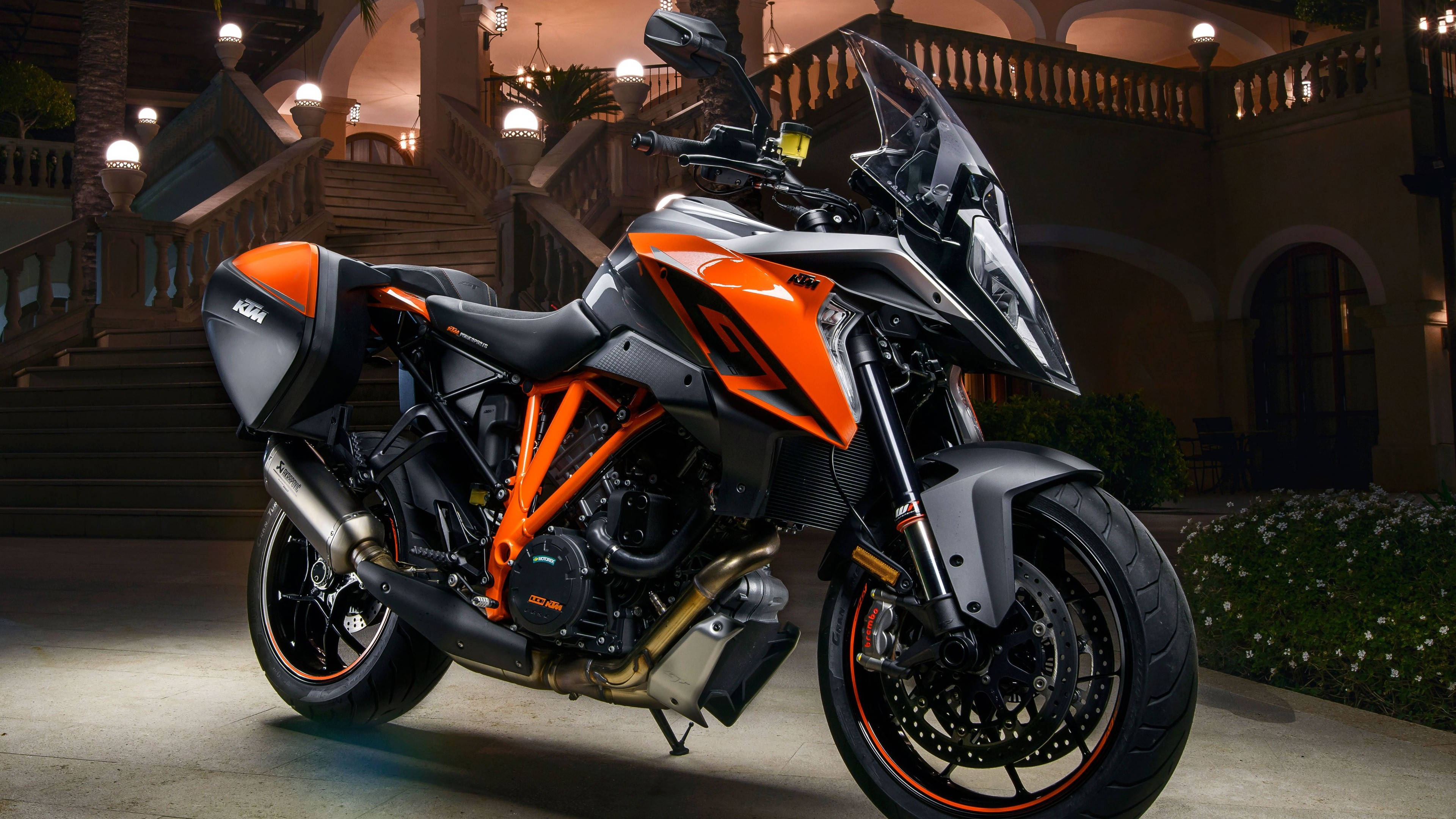 KTM 摩托車 桌布 壁紙 — 一輛時尚的橙色和黑色KTM摩托車在夜間停放在宏偉的石階上，周圍有溫暖的環境照明。