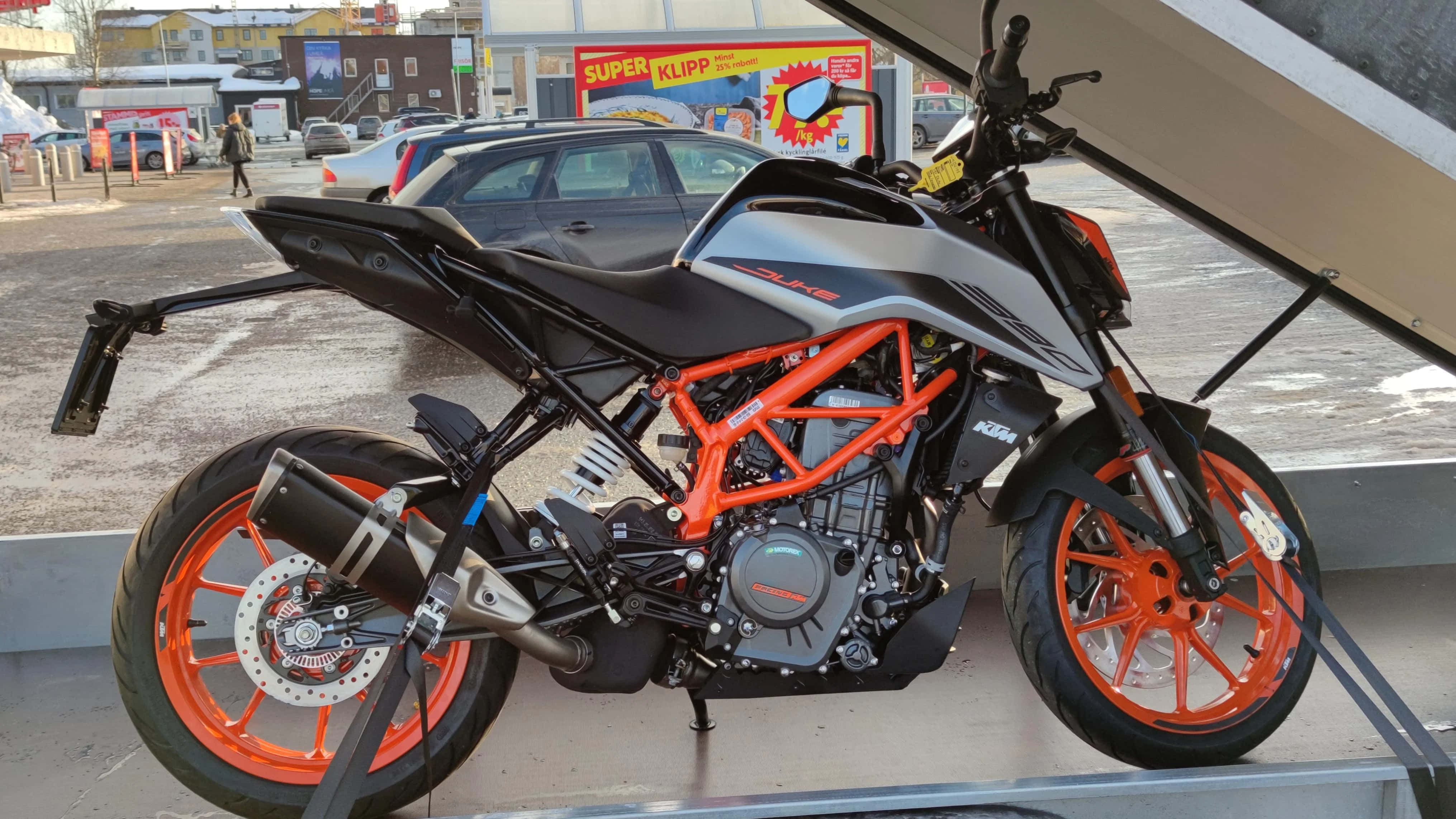 Ktm 桌布 壁紙 — 一輛時尚的橙色和黑色KTM機車停泊在 shelter 下，背景是城市建築和超市招牌。