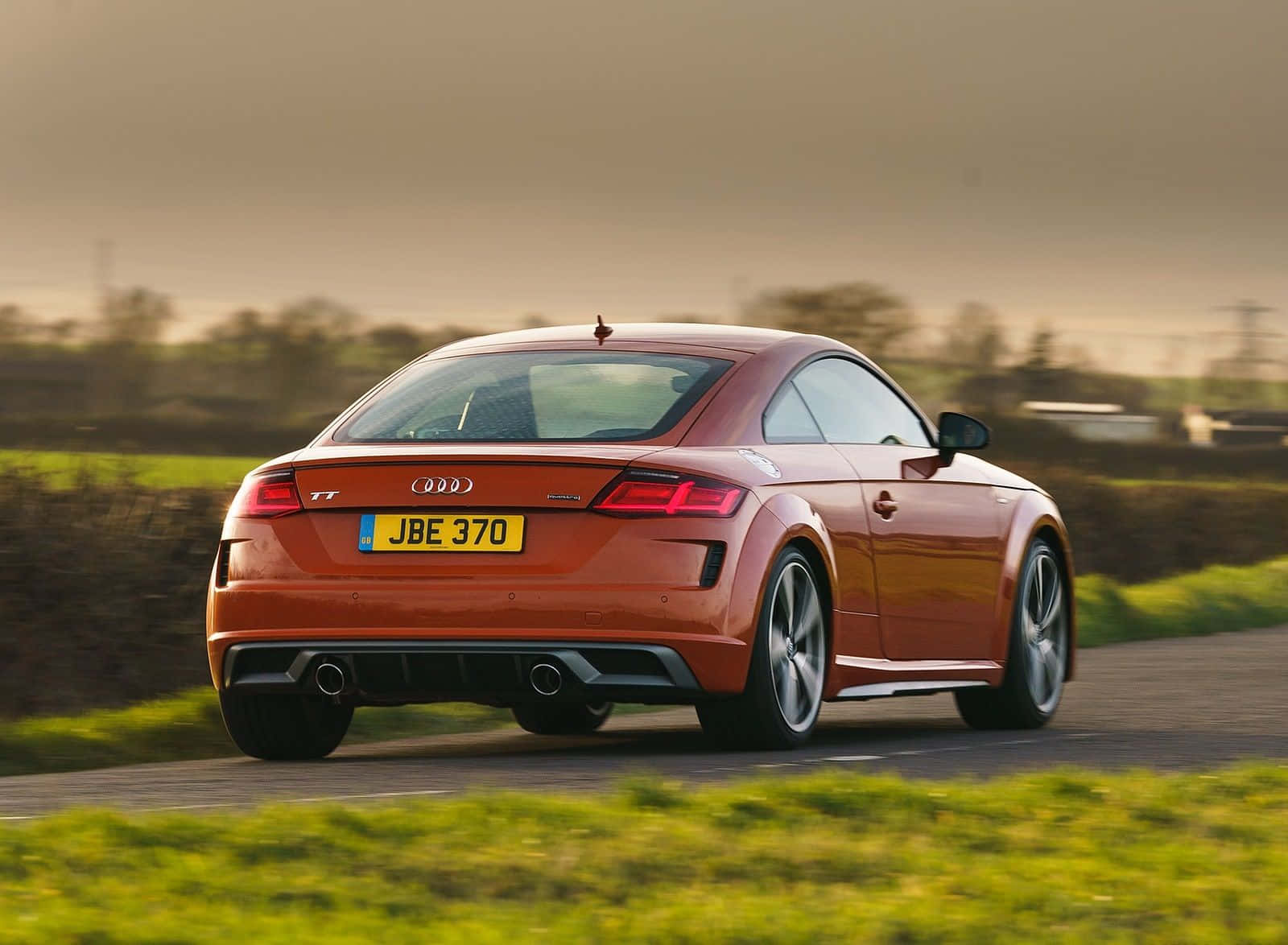 Audi Tt 桌布 壁紙 — 一輛時尚的橙色奧迪TT跑車在鄉村道路上行駛，背景是模糊的綠色田野和樹籬，天空柔和金黃。