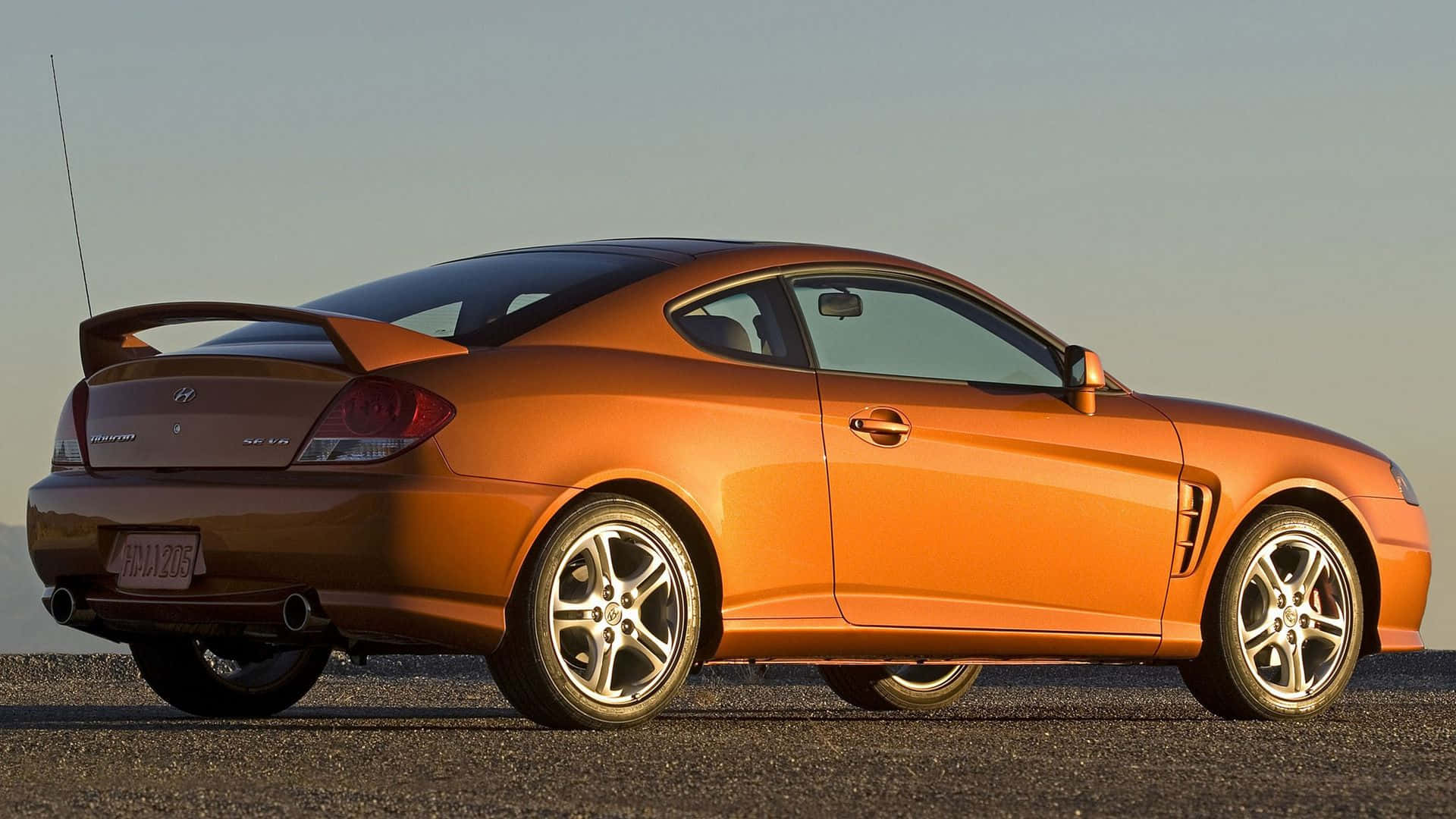 Hyundai Tiburon 桌布 壁紙 — 一輛時尚的橙色跑車，黑色車頂和銀色輪圈，停在晴朗天空下的瀝青路面上。
