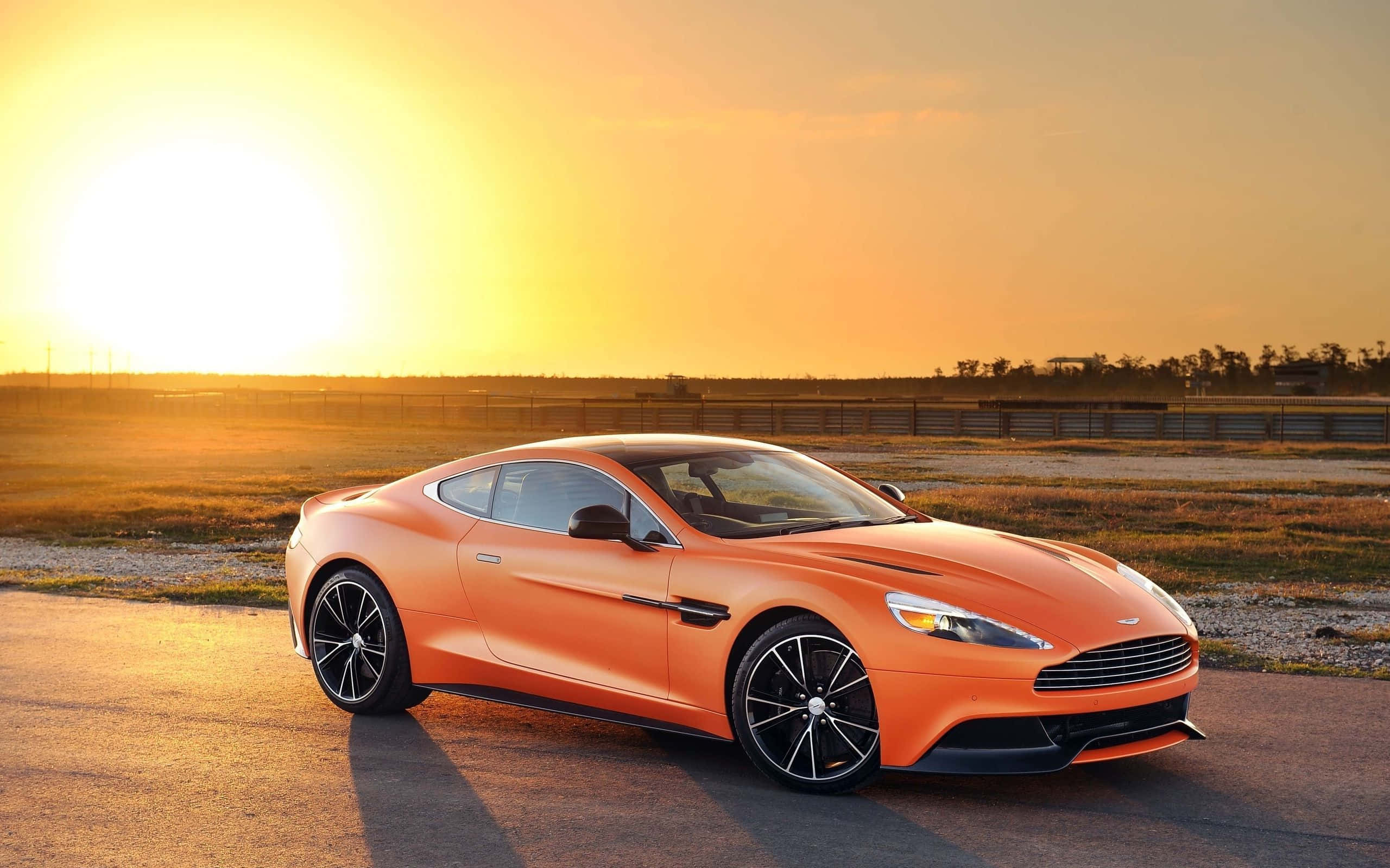 Aston Martin Vanquish 桌布 壁紙 — 一輛時尚的橙色跑車在金色夕陽下閃耀，在開闊的鋪砌道路上投下長長的影子。
