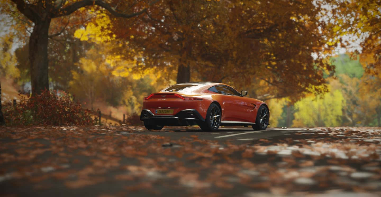 Aston Martin Vantage 桌布 壁紙 — 一輛時尚的橙色跑車在金色秋樹的樹冠下行駛在一條落葉滿佈的道路上。