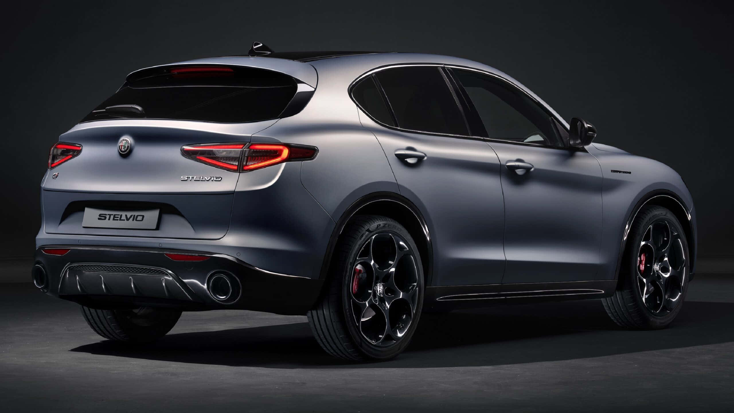 愛快羅密歐 桌布 壁紙 — 一輛時尚的消光灰色Alfa Romeo Stelvio SUV，從後方四分之三角度展示，帶有發光的紅色尾燈和黑色合金輪圈，背景為深色攝影棚。