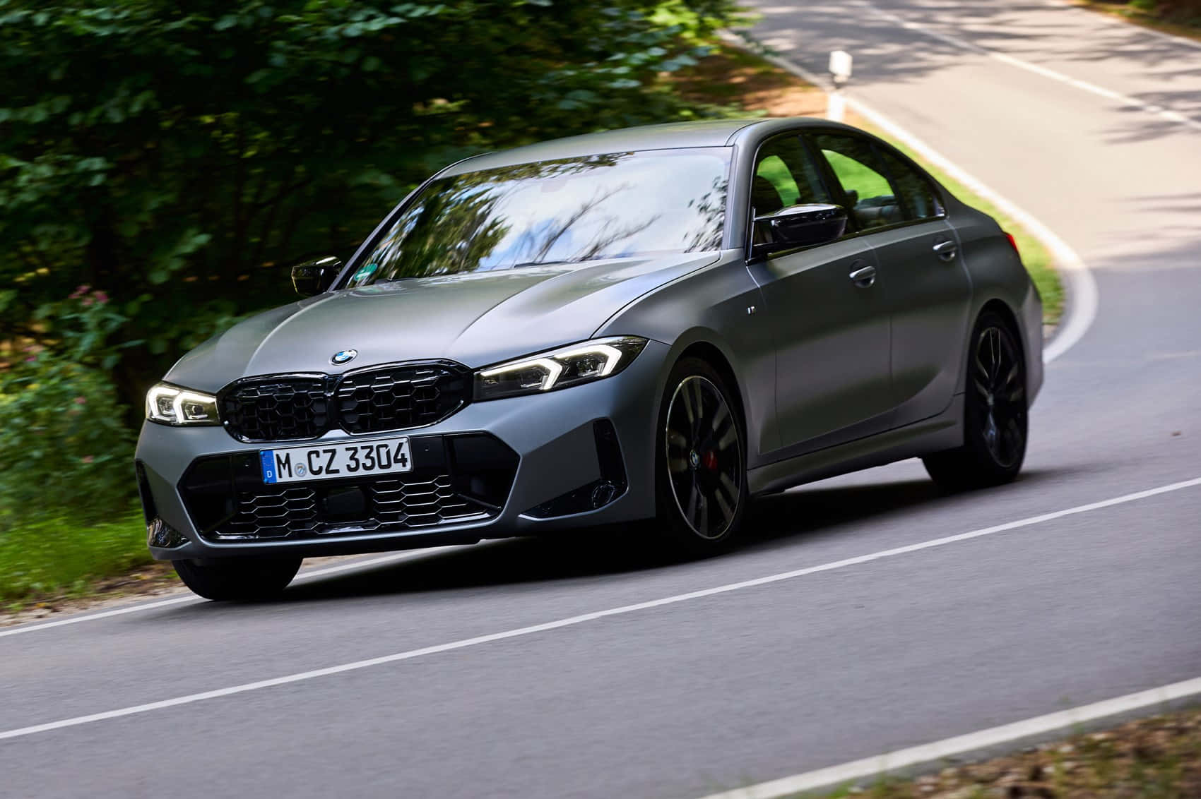 Bmw 3 Series 桌布 壁紙 — 一輛時尚的消光灰色BMW 3系列轎車在蜿蜒的森林道路上行駛，兩旁綠樹成蔭。