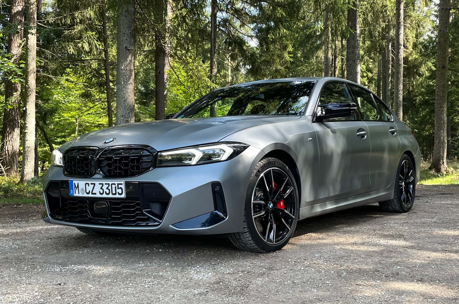 Bmw 3 Series 桌布 壁紙 — 一輛時尚的消光灰色BMW M3轎車，配備黑色輪圈和紅色制動卡鉗，停在陽光普照的松樹林碎石地上。