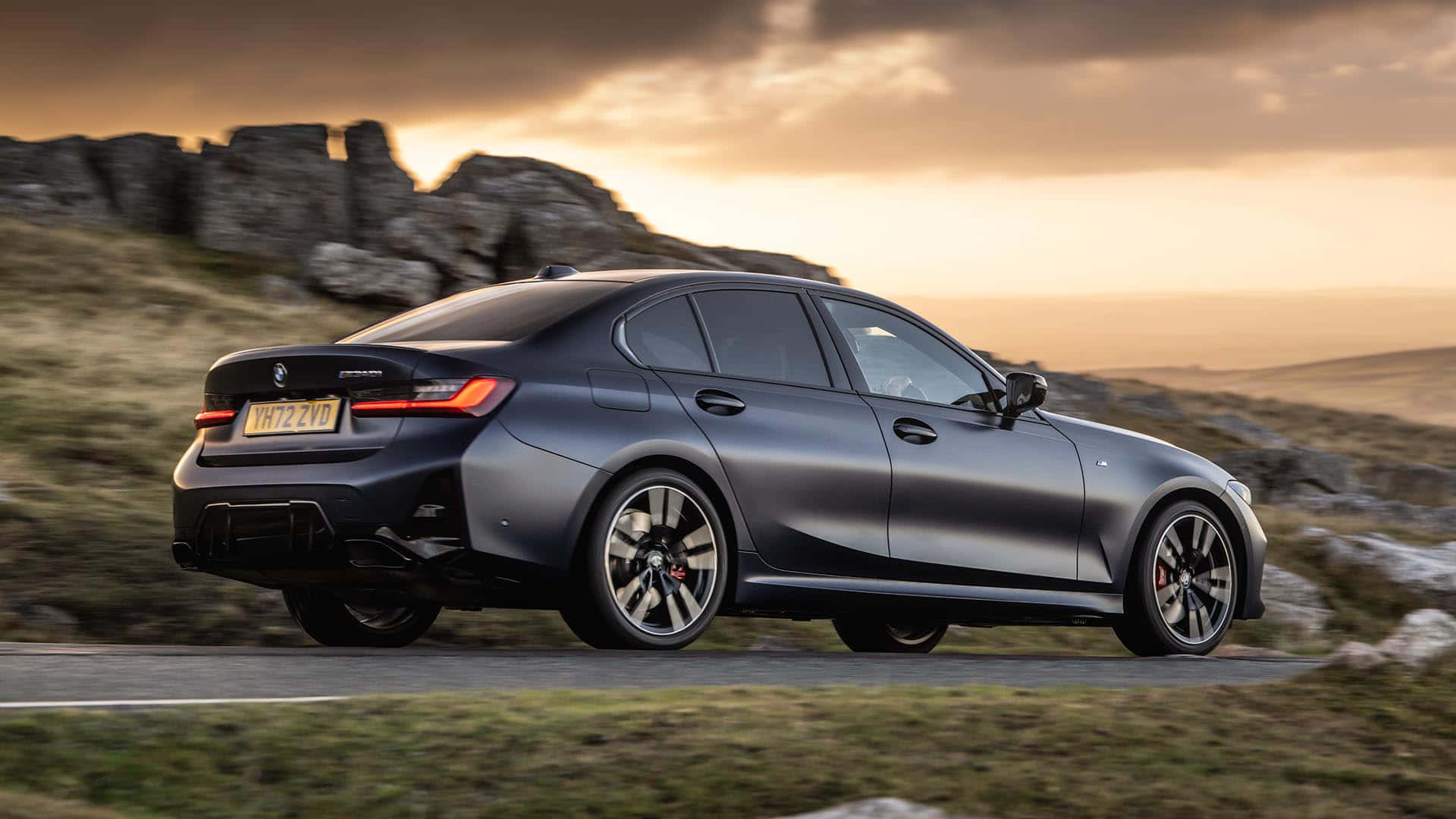 BMW 動圖 桌布 壁紙 — 一輛時尚的消光灰色BMW M3轎車在日落時分沿著蜿蜒的鄉村道路行駛，背景是岩石丘陵。