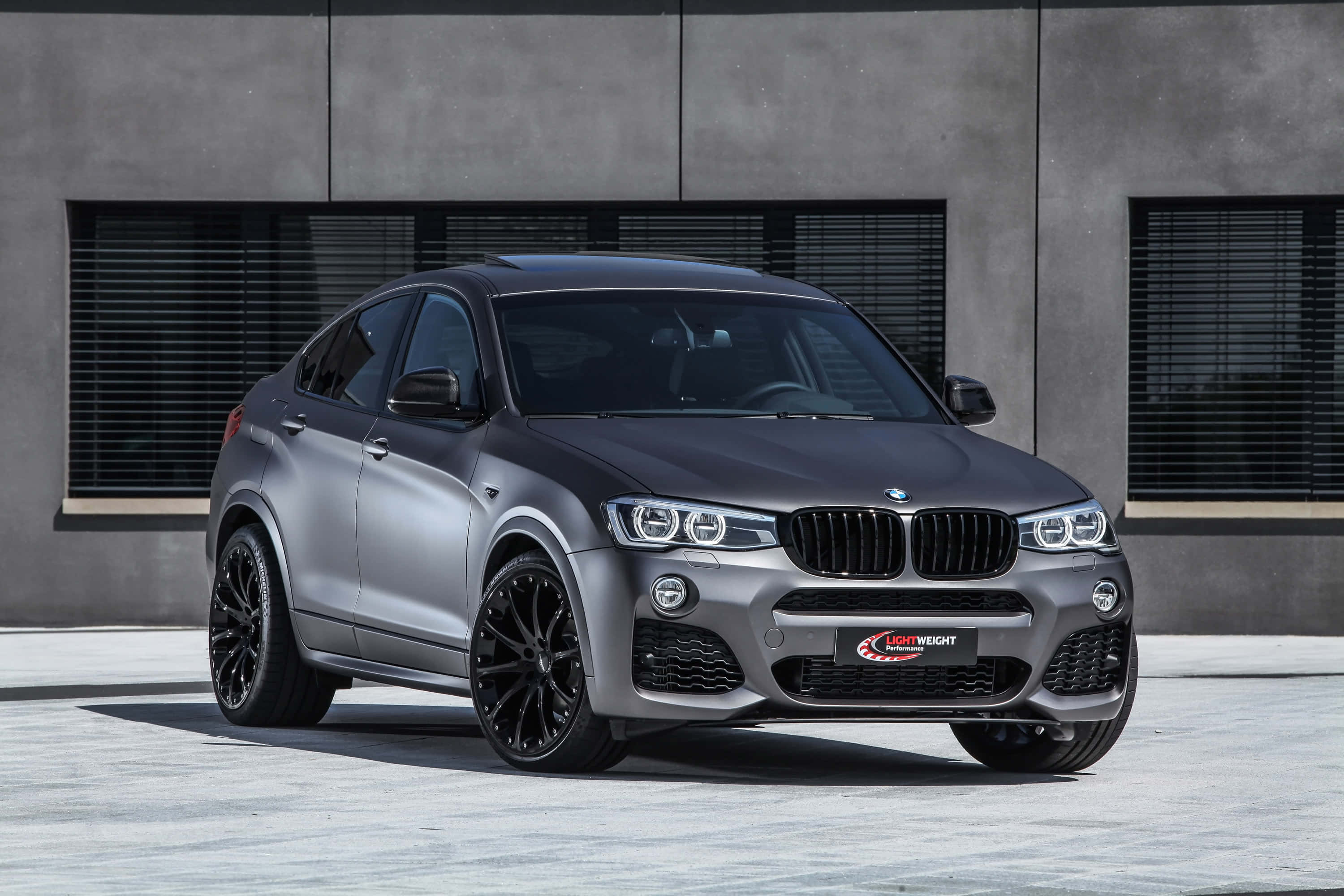 Bmw X4 桌布 壁紙 — 一輛時尚的消光灰色BMW X4搭配黑色輪圈，在明亮的日光下停在一棟現代混凝土建築前。