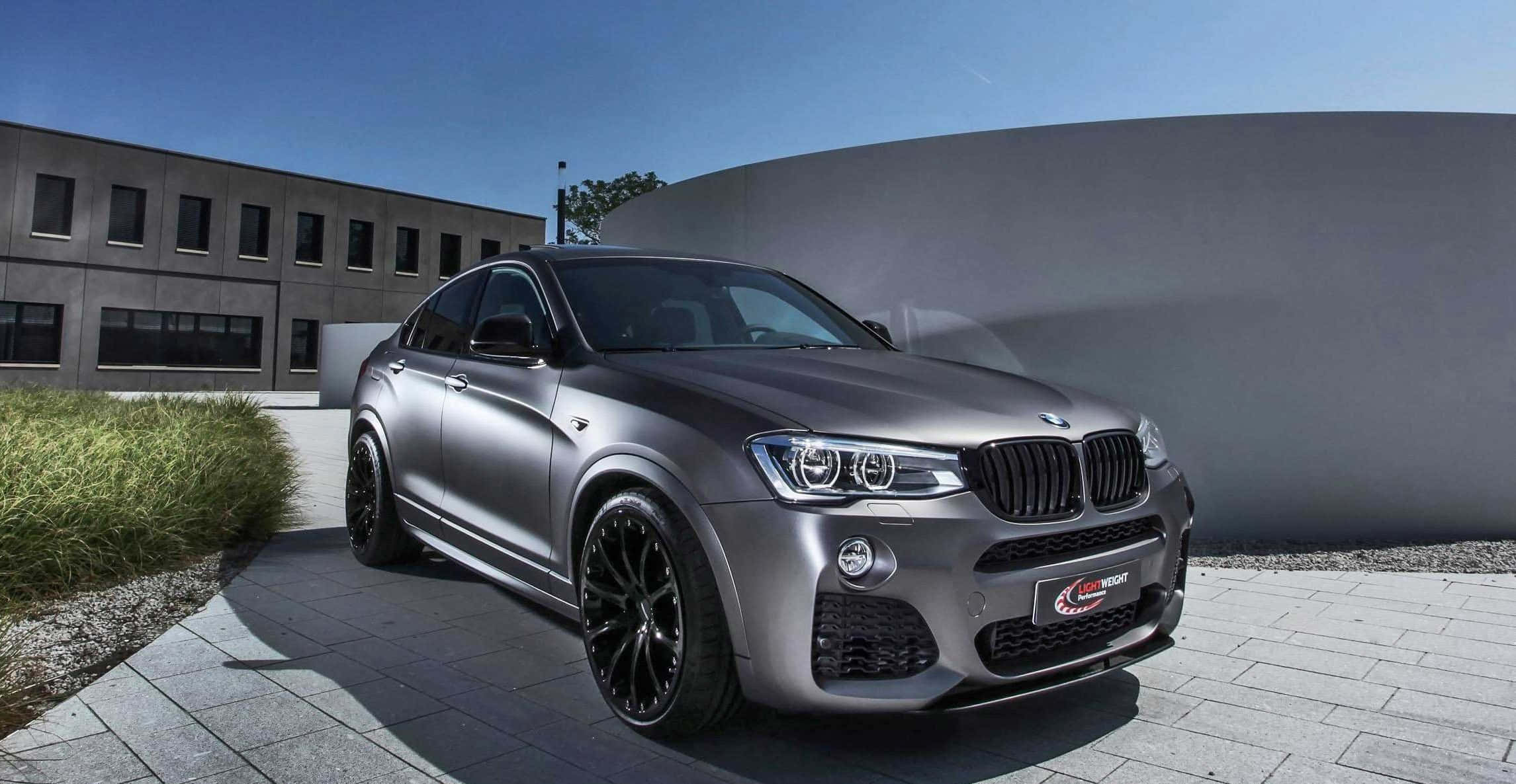 Bmw X4 桌布 壁紙 — 一輛時尚的消光灰色BMW X6M跑車停放在鋪設瓷磚的庭院中，周圍有現代建築和綠植，頭頂是清澈的藍天。