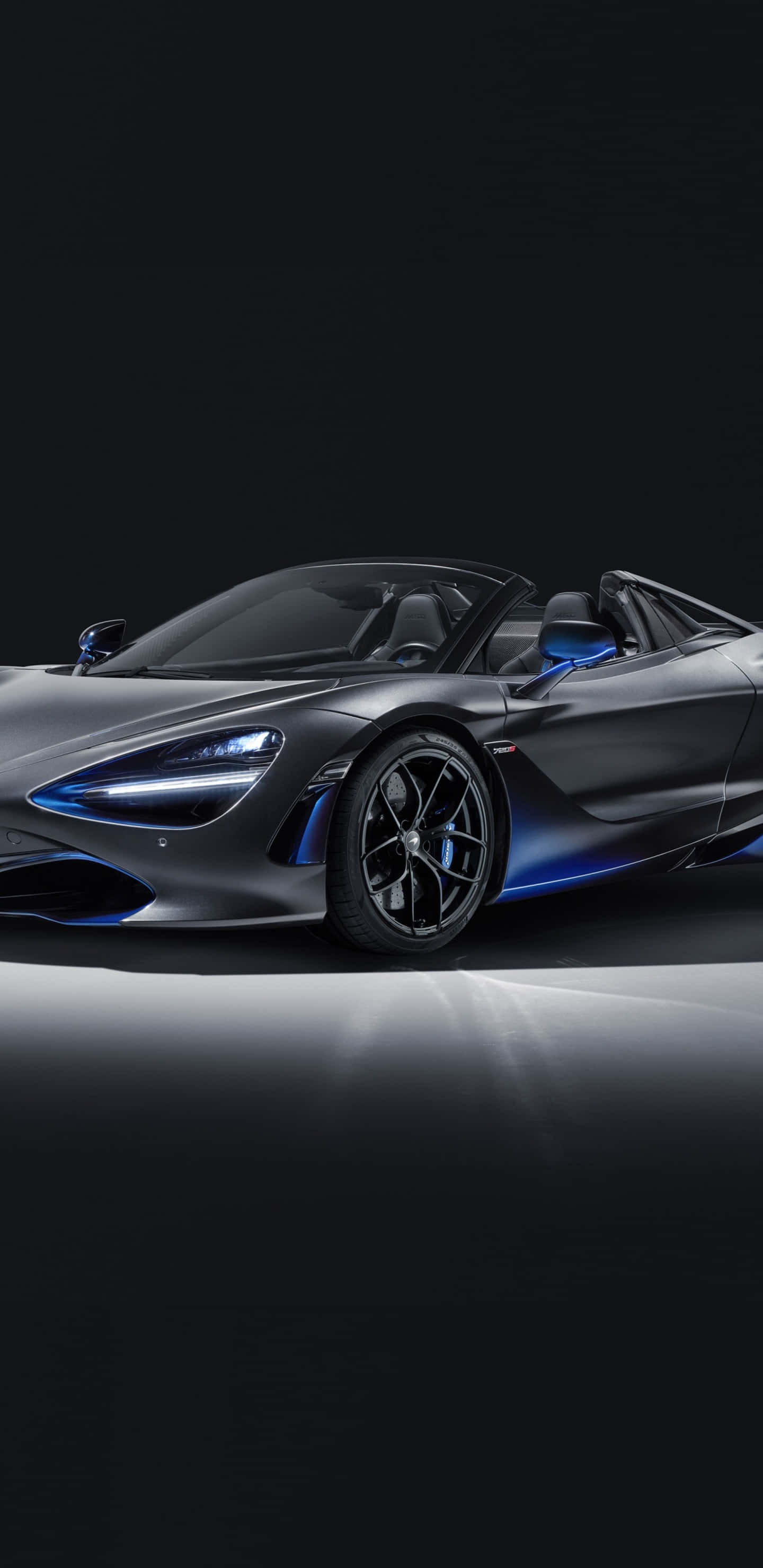 Pixel 3xl Mclaren 720s 桌布 壁紙 — 一輛時尚的消光灰色McLaren 720S Spider，帶有發光的藍色飾邊和車頭燈，在極簡主義工作室中以戲劇性的聚光燈展示。