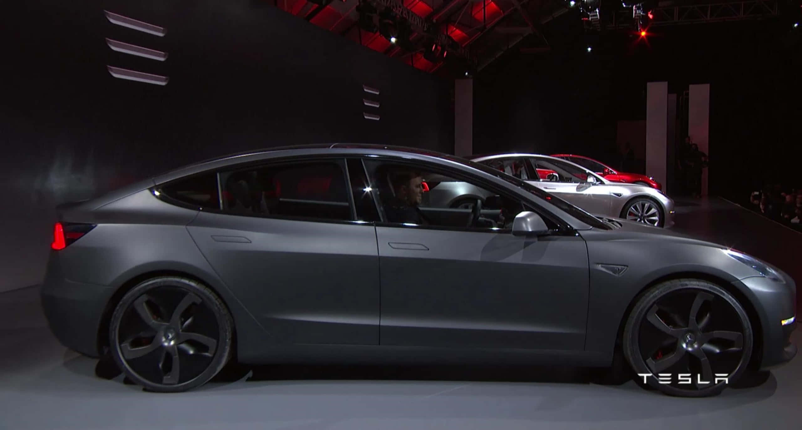Tesla Model 3 桌布 壁紙 — 一輛時尚的消光灰色Tesla Model 3在昏暗的舞台上展示，配有紅色重點照明，背景中可見另一輛車。