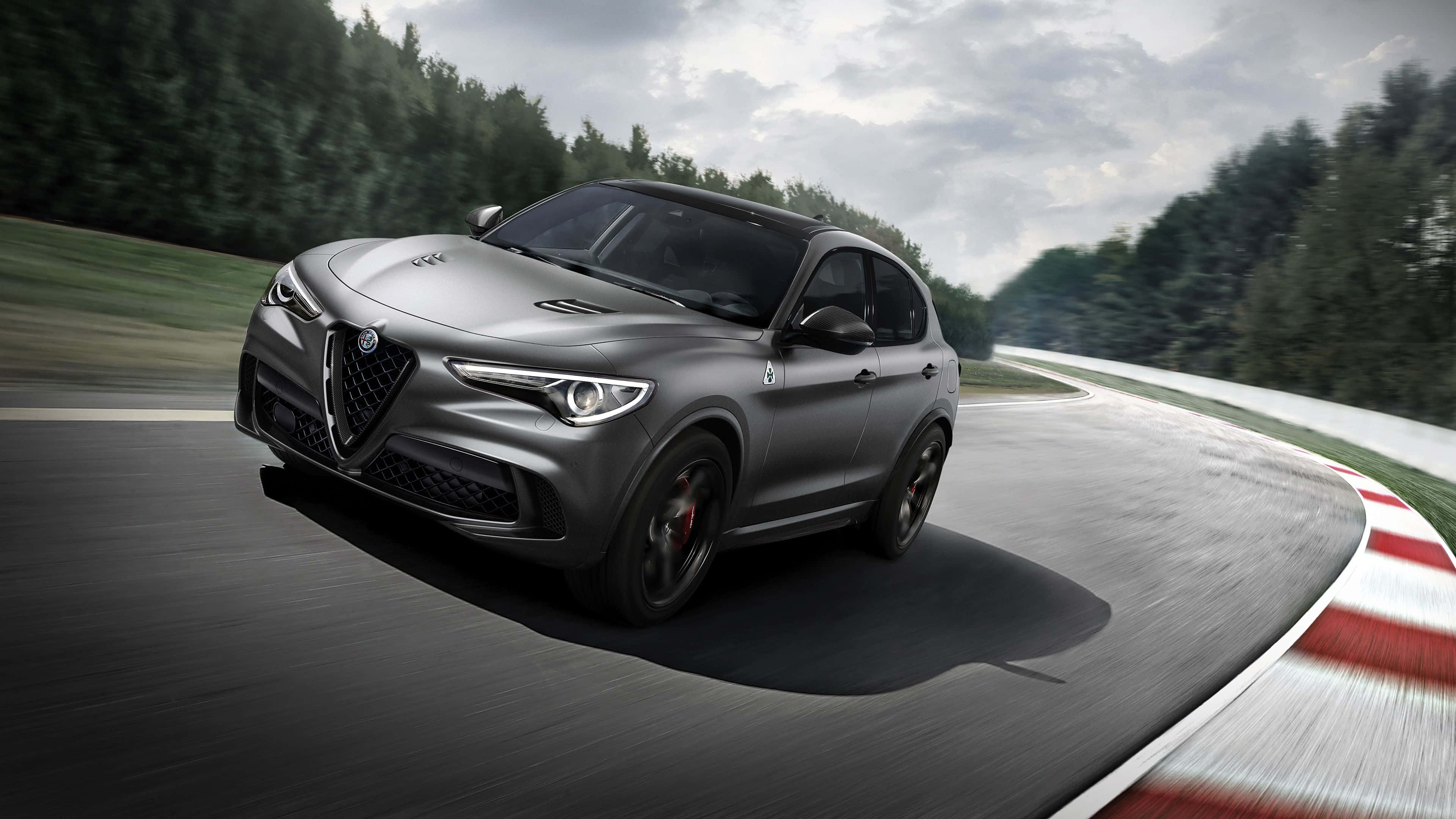 Alfa Romeo Stelvio 桌布 壁紙 — 一輛時尚的消光灰色愛快羅密歐SUV在陰天下一條蜿蜒的賽道上奔馳，賽道邊緣有紅白路緣石和茂密的綠色森林。