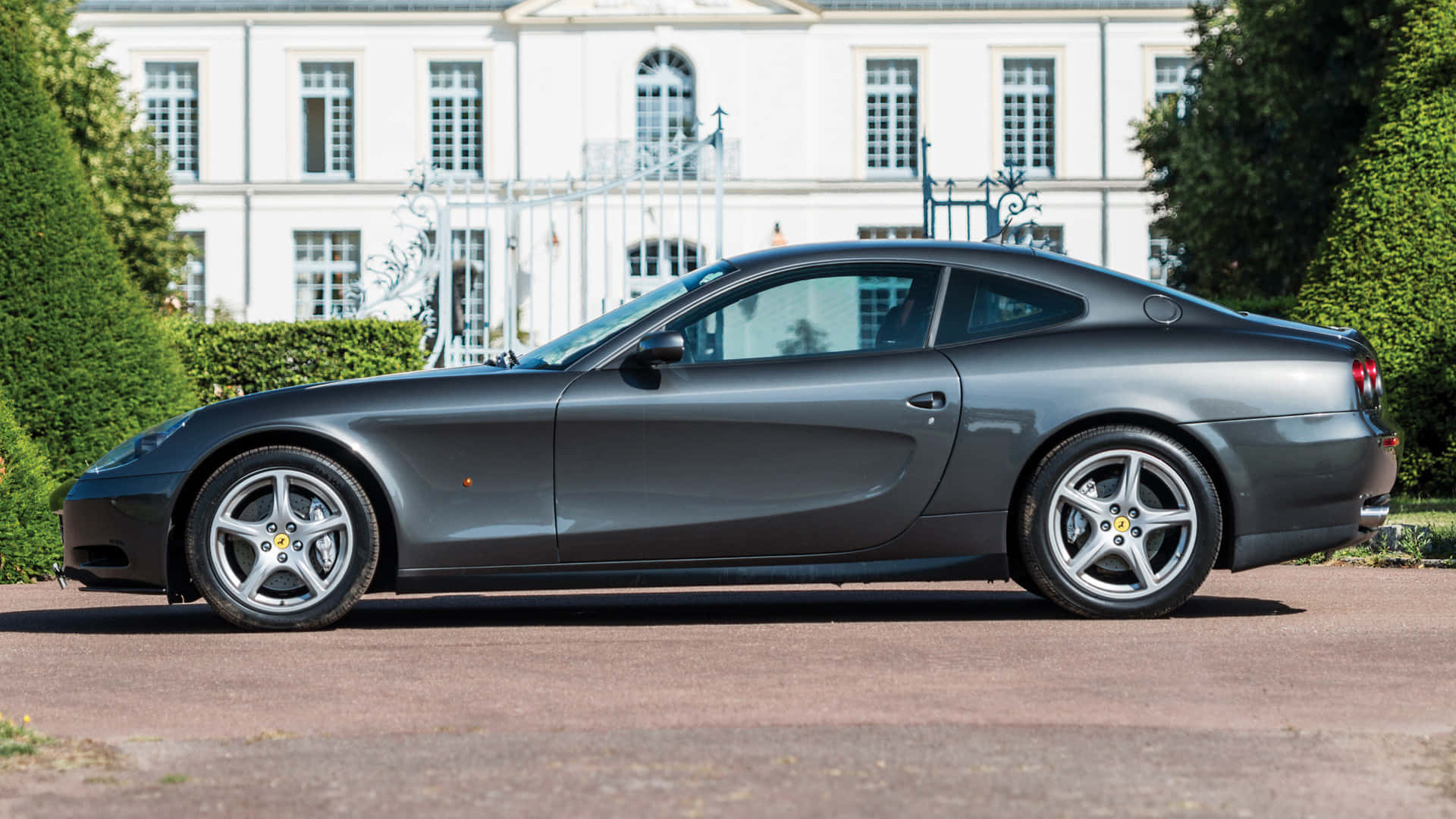 Ferrari 612 Scaglietti 桌布 壁紙 — 一輛時尚的消光灰色法拉利612 Superamerica停放在一座帶有修剪整齊樹籬的白色豪宅前的碎石車道上。