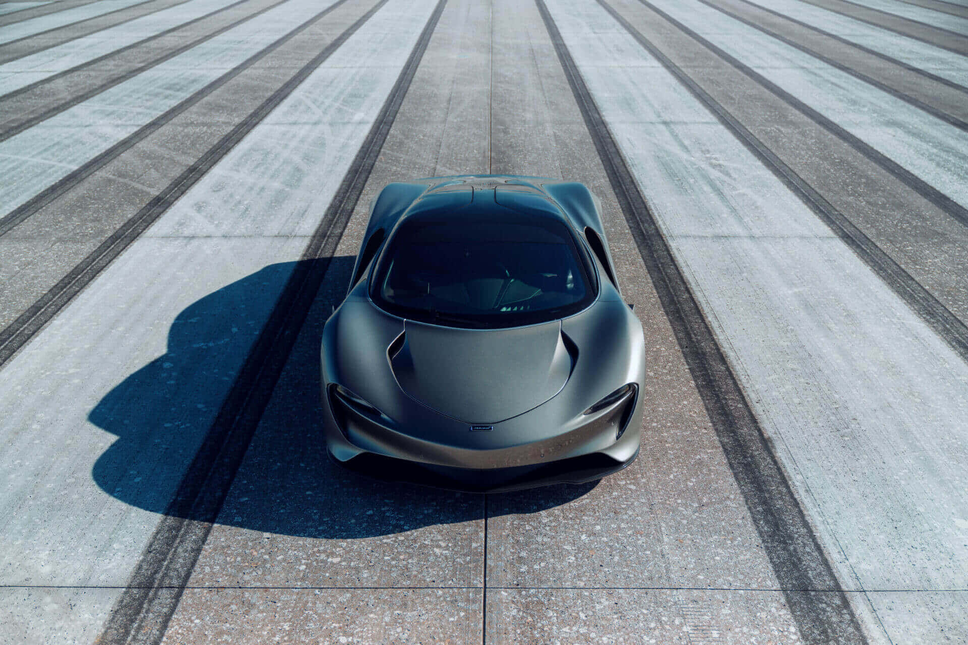 Mclaren Speedtail 桌布 壁紙 — 一輛時尚的消光灰色跑車在明亮的日光下，居中停放在一條帶有平行輪胎痕的混凝土賽道上。