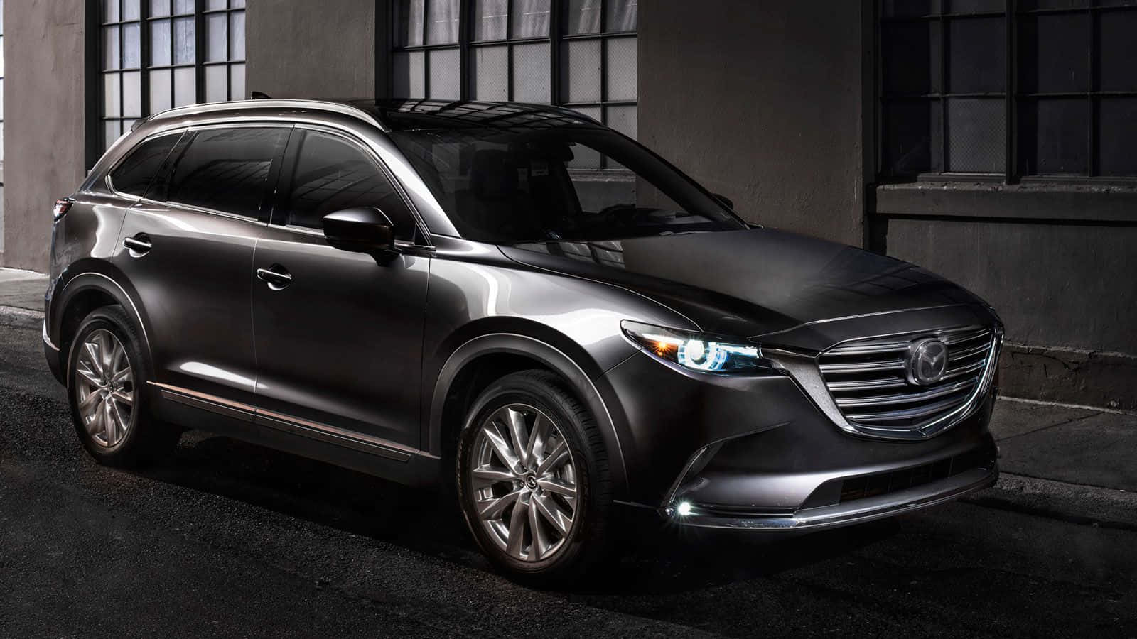 Mazda Cx-9 桌布 壁紙 — 一輛時尚的消光灰色馬自達CX-9 SUV在夜間停在一棟有大窗戶的建築物前。