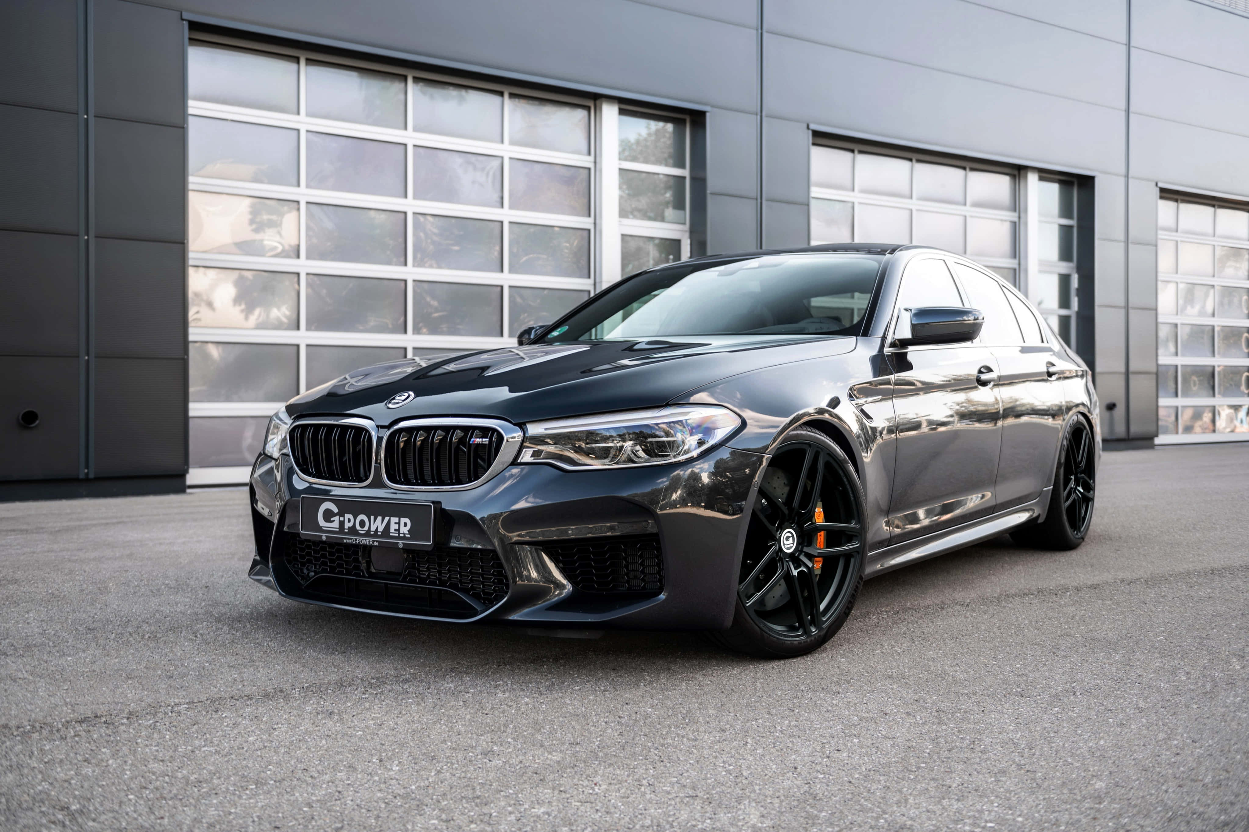 桌布 壁紙 — 一輛時尚的消光黑色BMW M5，帶有橙色制動卡鉗，停在現代工業建築前。